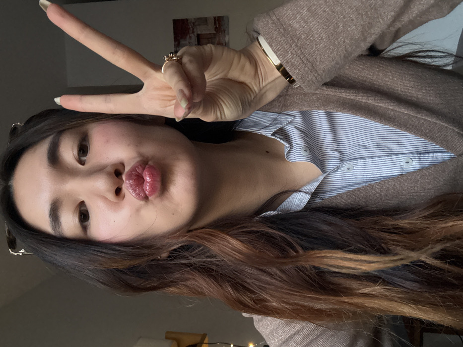 A female individual of East Asian descent is depicted in a chest-up, indoor photograph taken in the city of Paris, France. She has long, wavy dark brown hair with lighter, possibly auburn, highlights, and wears glasses pushed onto the top of her head. Her lips are puckered into a "kiss" pose, and her right hand is raised to the right of her face, displaying a "V" or peace sign with the index and middle fingers extended. She wears a light blue and white pinstriped collared shirt beneath a V-neck tan cardigan. A gold-toned ring with a dark stone is present on her right ring finger, and a gold-toned bracelet with a white insert adorns her right wrist. The background features a light-colored wall. On the left, partial view of a wooden object and string lights are visible. On the right, a framed artwork depicting an aged building facade, possibly stone or brick, hangs on the wall.