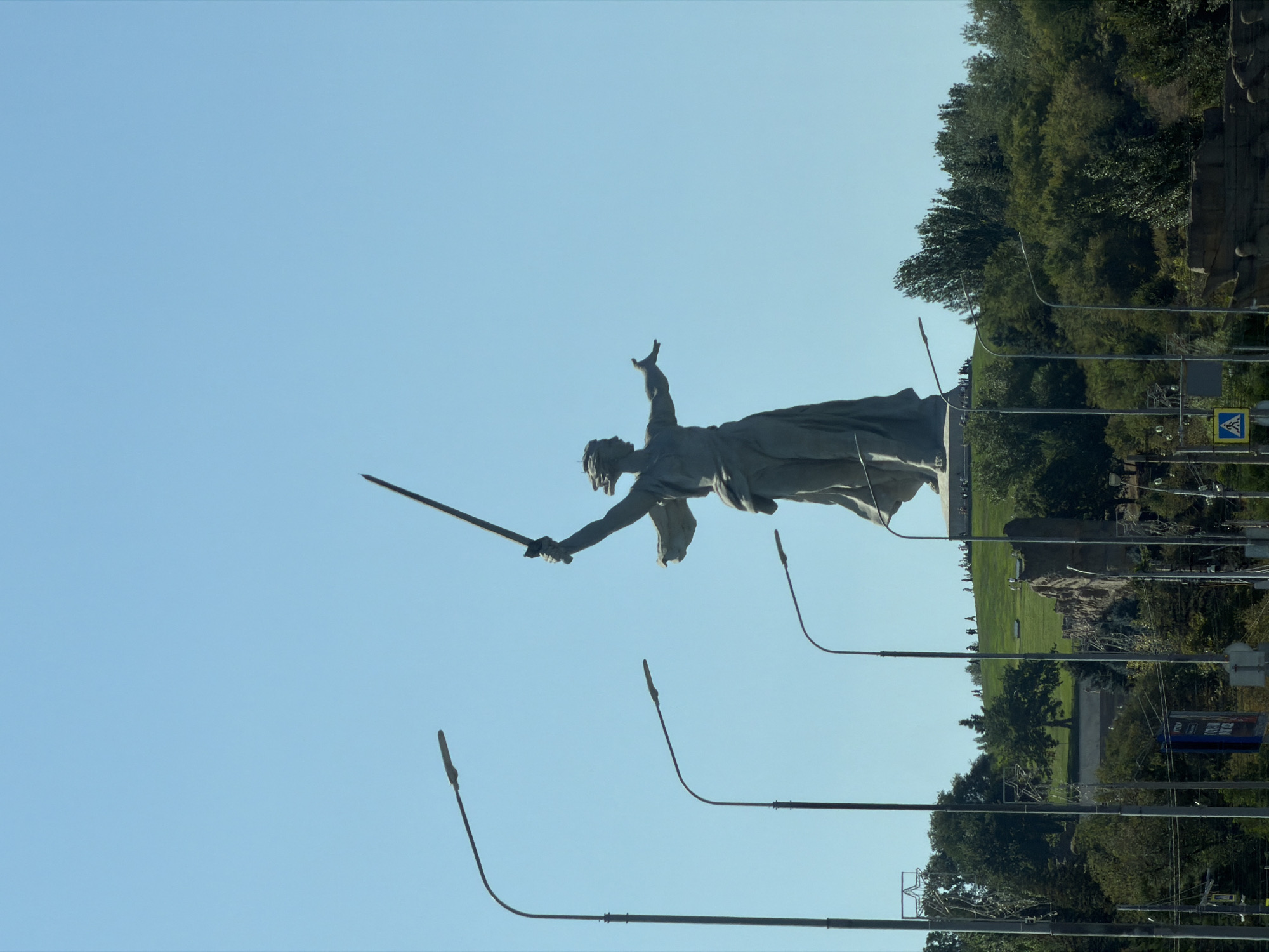 The image shows a view of the Motherland Calls monument, a colossal statue on Mamayev Kurgan in Volgograd, Russia. The monument depicts a woman with a raised sword, symbolizing the call to arms. The statue is made of concrete and stands on a hill covered with greenery and scattered rocks. Several streetlights are visible in the foreground, extending diagonally across the frame, with their lamps pointed downwards. In the background, a clear blue sky dominates the upper half of the image. Below the monument, there are a few small figures of people walking on the hill. The base of the monument is partially visible. Trees and foliage surround the base of the hill. A road or pathway is visible at the bottom, with some vehicles and signage, suggesting an urban or semi-urban environment.