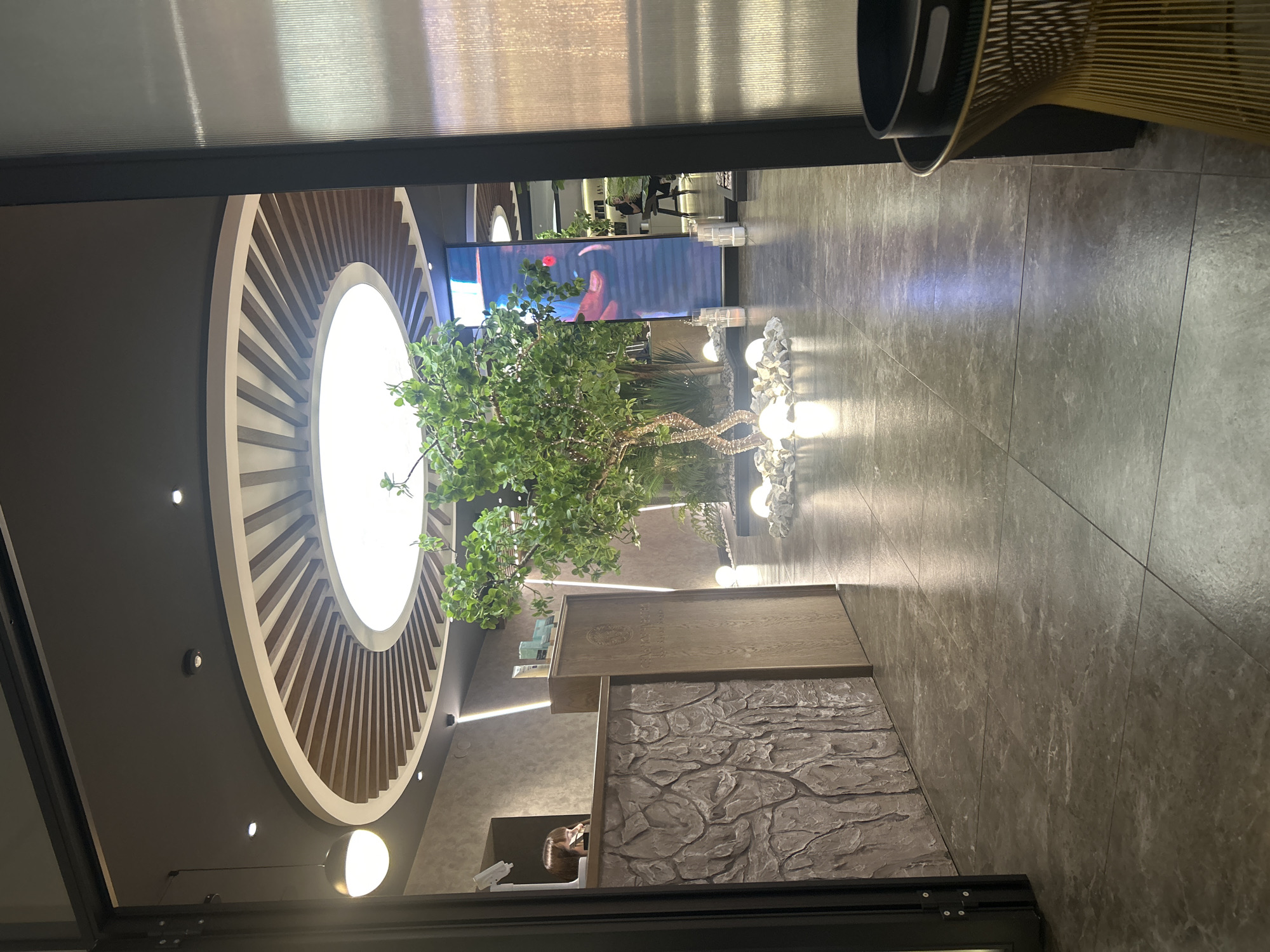 The image shows an interior view of a business or commercial space, likely a salon or spa.
Environment:
* The floor is covered in large, gray tiles.
* A circular ceiling feature with radial wooden slats frames a bright light.
* The walls are gray with recessed lighting.
* A desk with a faux rock texture on the front and a light wood-grained top is prominent.
Objects:
* A decorative tree with green foliage stands near the desk, surrounded by rocks and white spherical lights.
* A large digital display (possibly a TV) is visible in the background, showing what appears to be a person’s face.
* A chair with a woven back is partially visible in the upper right corner.
* There are hair products and other salon-related items visible on the desk and behind it.
Actions/Interactions:
* A person can be seen behind the desk, presumably working.
Inferred Location:
* The presence of specific products, signage, or design elements might imply the specific business. There is no way to determine the location in the city of Votkinsk, Russia, from the image alone.
