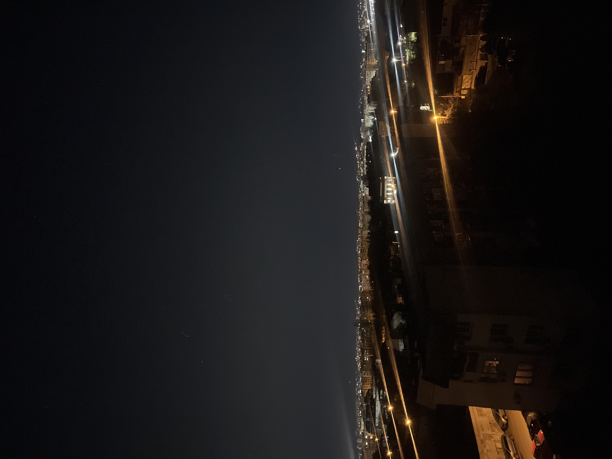 The image shows a nighttime urban landscape, likely in Sofia, Bulgaria, based on architectural styles and general layout. On the right side, illuminated residential buildings are visible, with parked cars and streetlights casting warm light onto the streets. The left side of the image is dominated by a dark sky with a few visible stars. A line of city lights stretches across the horizon, marking the extent of the urban area. The image gives the impression of an elevated perspective, as if the photograph was taken from a high vantage point. There are no people visible in the photograph. The focus is on the urban environment and its illumination at night.
