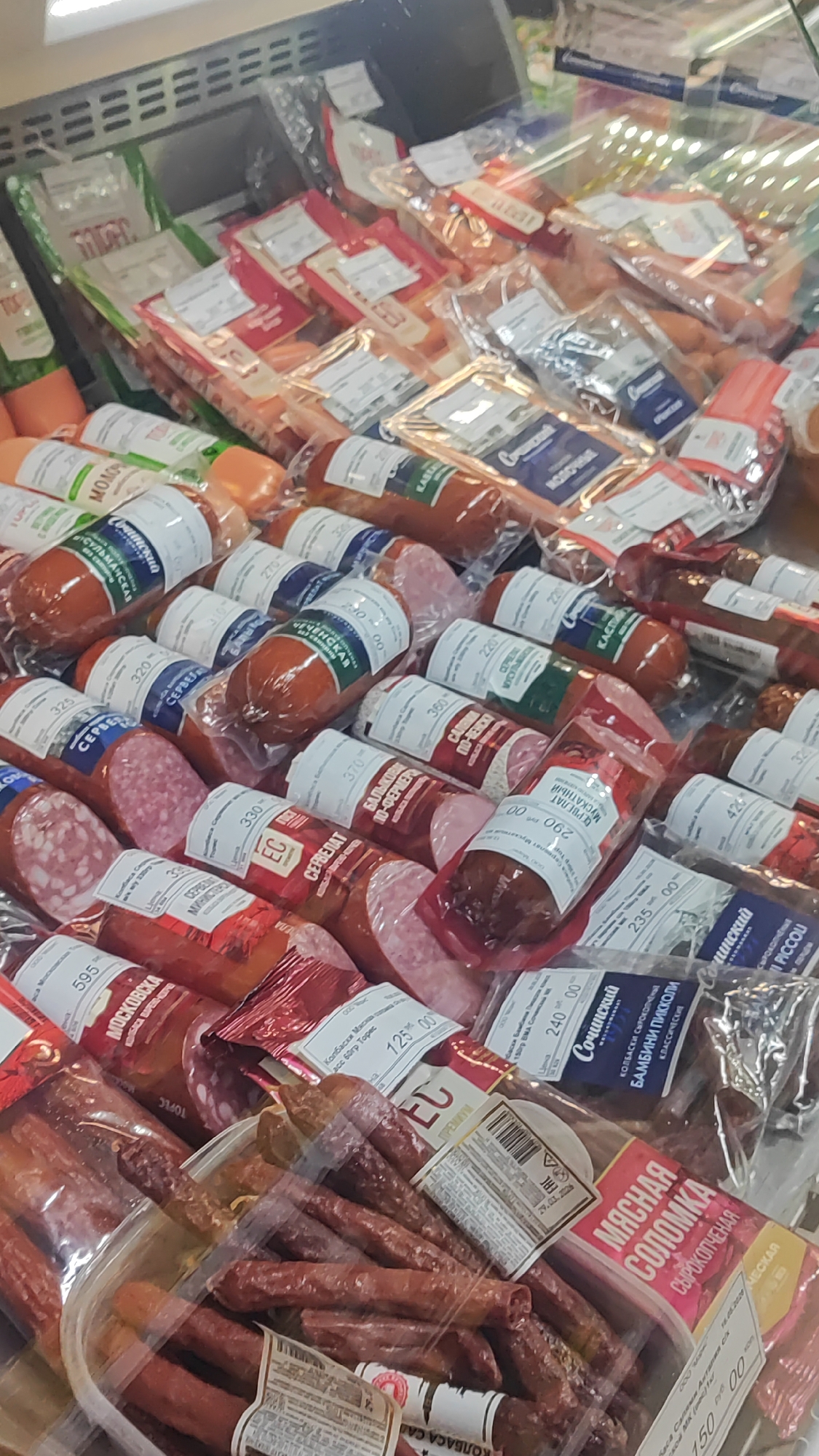 The image shows a refrigerated display case filled with a variety of packaged sausages. The display case appears to be located in a supermarket or grocery store in Sochi, Russia, as indicated by the Cyrillic text on the product labels, which includes brand names and product descriptions in Russian. The sausages are of various shapes, sizes, and packaging types, including round, cylindrical, and smaller, stick-like varieties. Some packages are transparent, allowing the contents to be clearly visible, while others have printed labels. The lighting suggests an indoor commercial environment. No people are visible in the image.