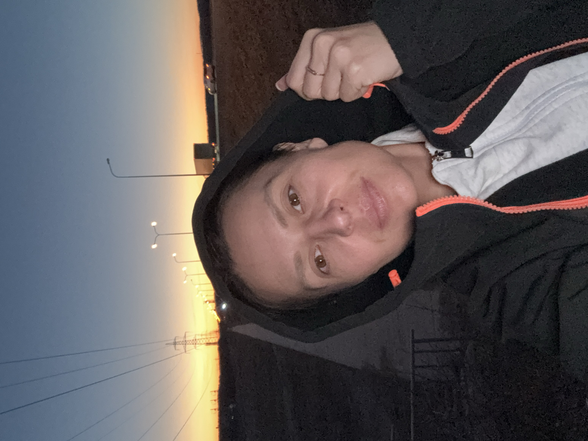 A vertically-oriented photograph shows a woman wearing a black hooded jacket with an orange zipper trim over a white shirt. She is looking at the camera. Her left hand is visible holding the collar of the hood. The background shows a sunset with a gradient from orange to blue. Several lampposts and power lines are visible. The location is Chaikovsky, Russia.