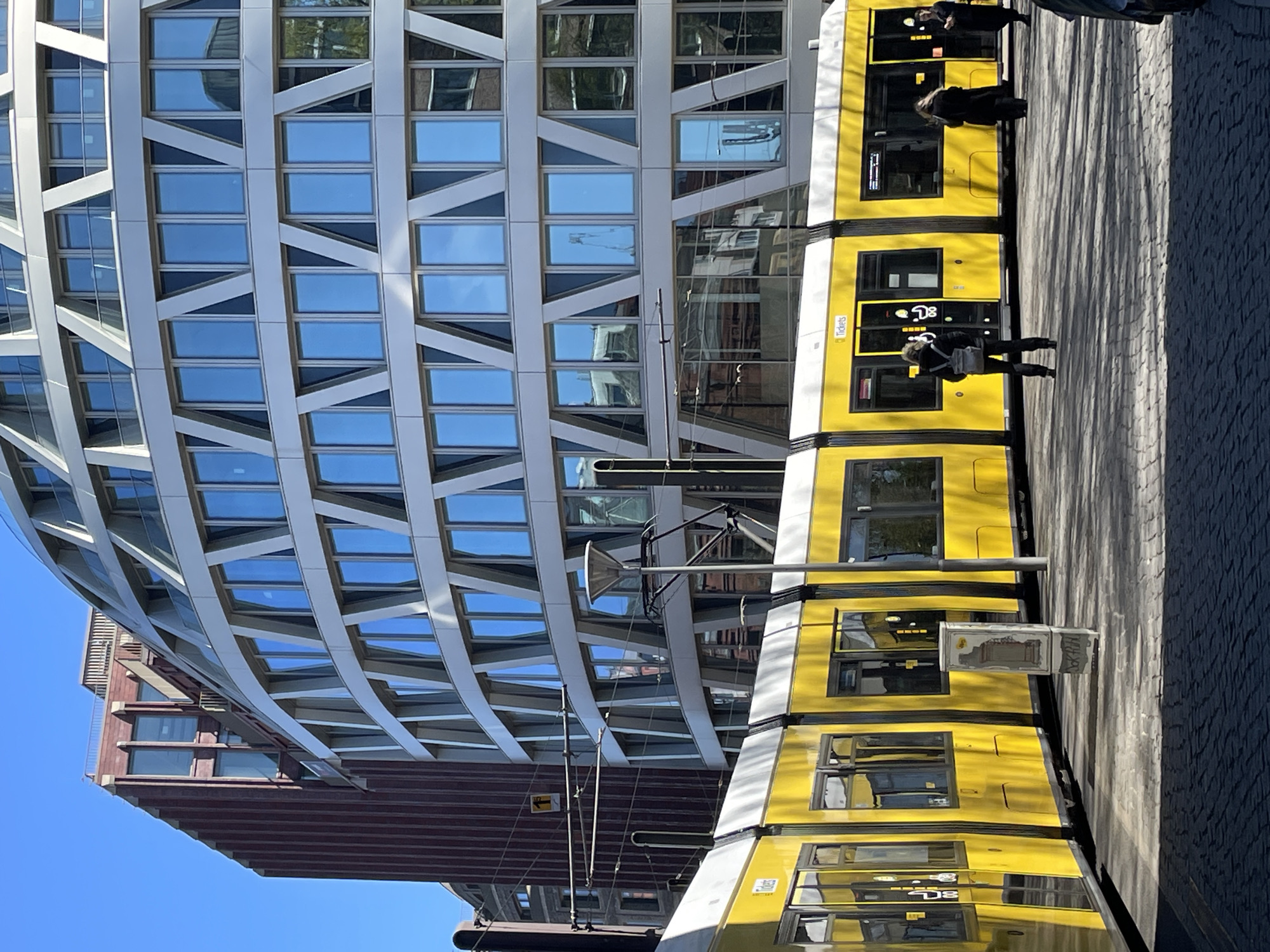 The image presents a bright, urban street scene in Berlin on a clear and sunny day. A striking yellow tram, characteristic of the city's public transportation, occupies a significant portion of the right side of the frame, appearing to be stopped or in motion on its tracks.
On the left, a modern building features a distinctive, curved facade. This facade is characterized by a geometric grid of light-colored supports creating a pattern of trapezoids and rectangles, interspersed with numerous windows. These windows reflect the bright blue sky and glimpses of the surrounding architecture. Behind this contemporary structure, part of an older, reddish-brown building with horizontal window lines is visible against the clear sky.
Several people are observed on the paved ground next to the tram. They appear to be adults, standing or walking, possibly waiting for or leaving the tram. One person is visible near the front section of the tram, and another further along its length.
The tram itself is predominantly bright yellow with white accents. Visible text on its side includes "Tickets" near a door, and a route or line indicator displaying "D8". Overhead electrical lines, part of the tram's power supply system, are also visible.
The ground is paved with dark gray cobblestones or similar material. Long shadows are cast across the pavement, suggesting the sun is not directly overhead, indicating either morning or late afternoon. A small, white utility box or newsstand is positioned close to the tram tracks, with discernible text including "NORLYK" and "ZEITUNG" (German for newspaper). The overall scene captures a typical, lively day in the German capital.