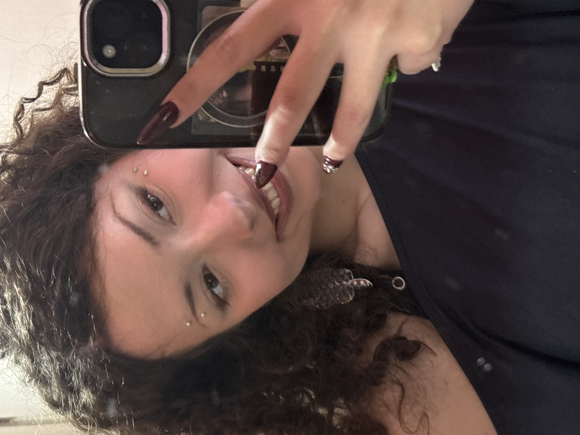 A person with curly brown hair is holding a smartphone in front of their face, partially obscuring it. The phone has a dual-lens camera and a dark case. The person has fair skin, brown eyes, and is wearing two small, round piercings above their left eyebrow. They are wearing a dark-colored top. The person's hand with long, dark red nails decorated with rhinestones on one finger is visible. The background is out of focus and appears to be a light, neutral color. There is no indication that this image was taken in Milan, Italy.