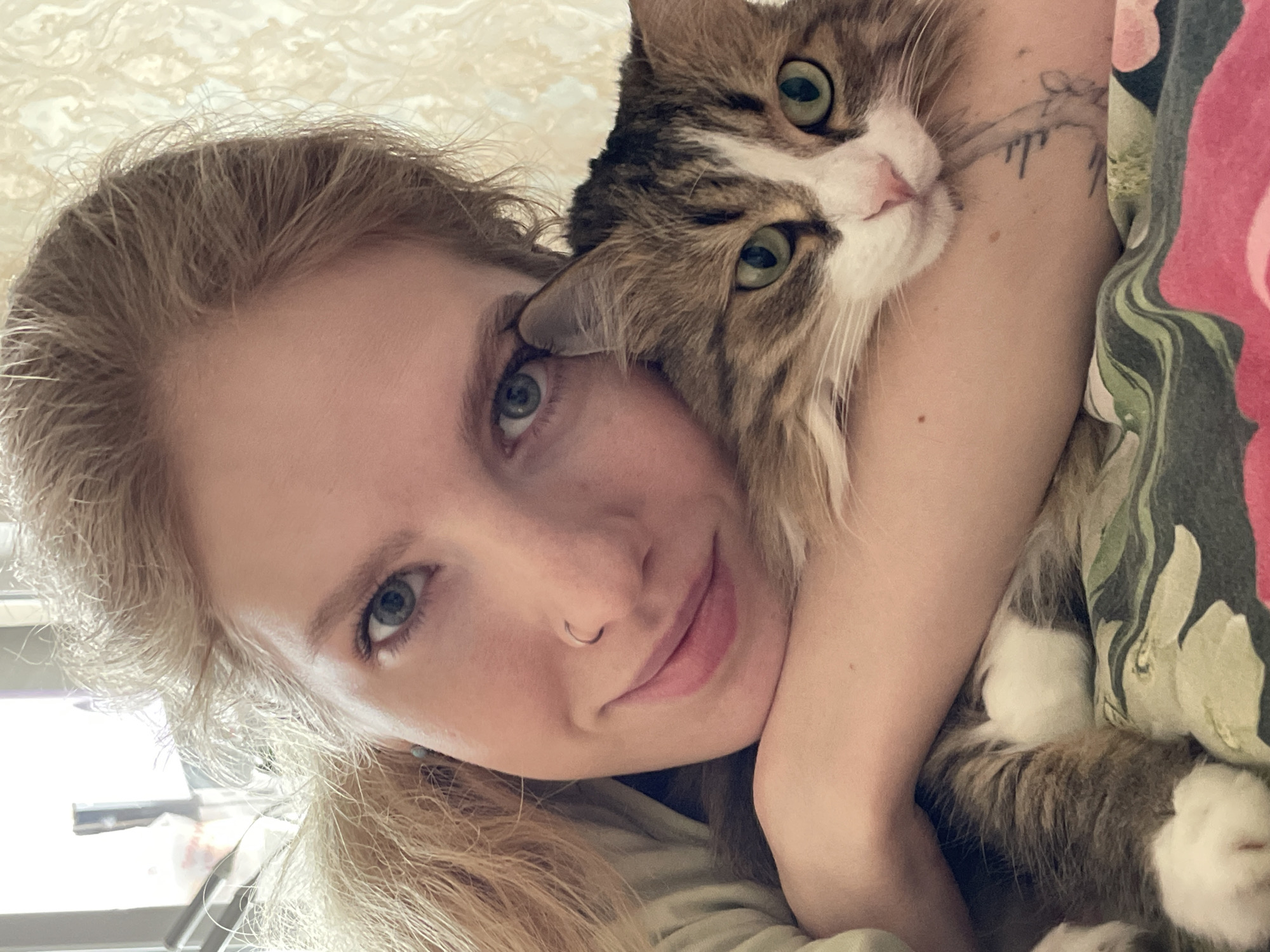 A woman is holding a cat. The woman has blonde hair, blue eyes, and a nose piercing. The cat is a brown and white tabby with green eyes. The woman's arm is around the cat's body, and the cat's head is resting on the woman's shoulder. The woman is smiling. The background is blurry, but appears to be a room with a window. There is a floral pattern on fabric next to the woman's arm. There is a tattoo on her arm near her elbow. The location in the city of Izhevsk, Russia cannot be determined.