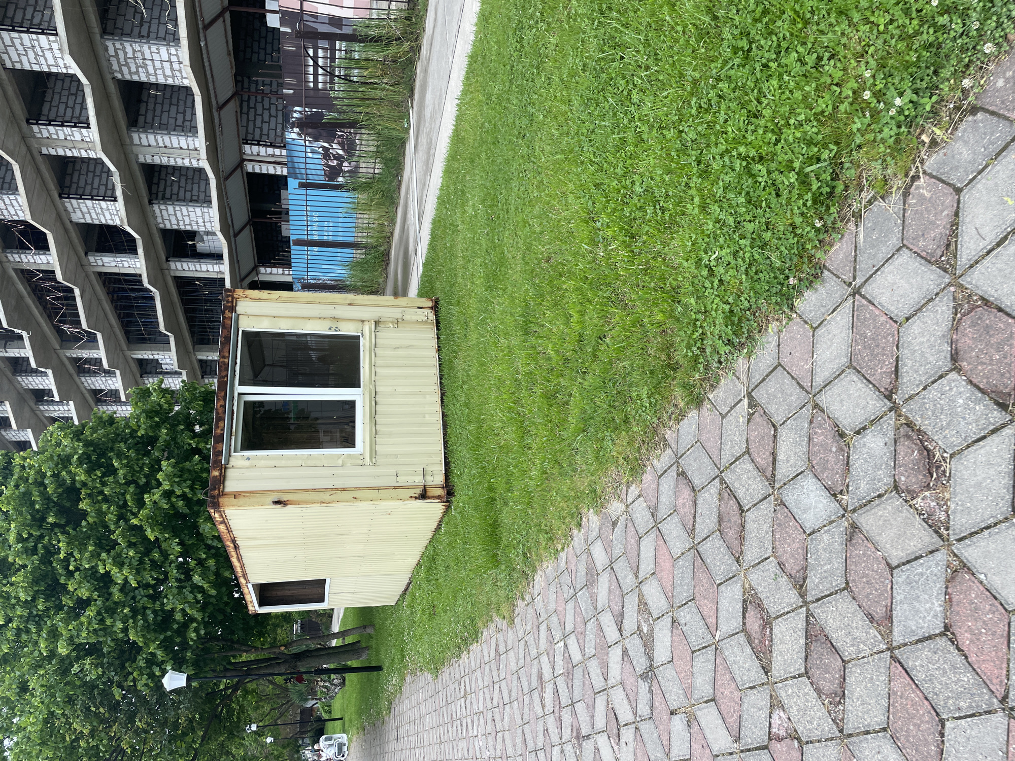 A medium shot, rotated approximately 90 degrees counter-clockwise, presents a paved walkway composed of interlocking gray and red geometric blocks. Adjacent to the walkway is a patch of green grass. A pale yellow, rectangular structure, potentially a modified shipping container, with two framed windows, sits partially on the grass and next to a building. To the left, a multi-story, unfinished concrete building is visible. In the distance, a street with parked cars and streetlights can be seen, implying an urban or suburban setting. The location is Olginka, Russia.