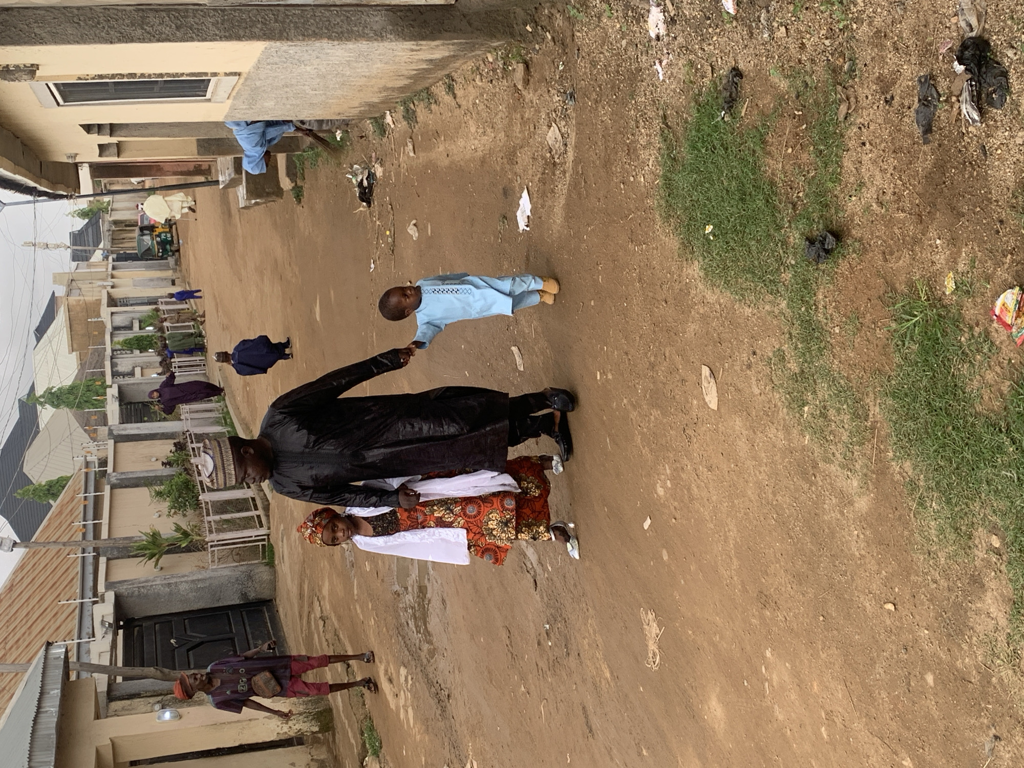 The image shows a dirt street in Jos, Nigeria. A man in a black robe holds hands with a child in a blue outfit. A woman in an orange and white dress walks beside the man. Other people walk further down the street. The street is lined with houses and buildings. The ground is mostly dirt with some patches of grass and litter.