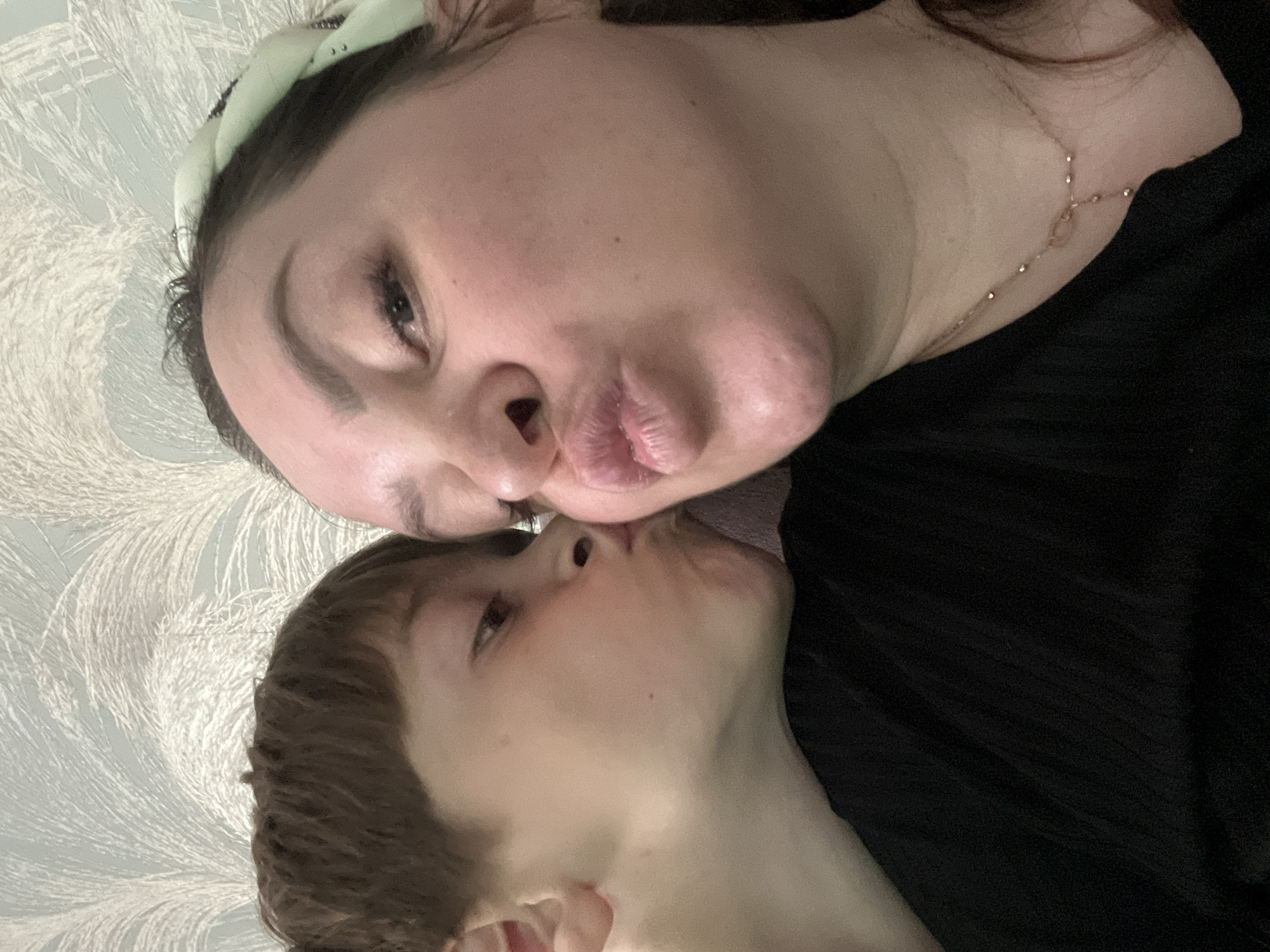 An adult woman and a child are depicted in a close-up, head-and-shoulders portrait, both puckering their lips in an affectionate gesture. The scene is located in Noviy, Russia.
The woman, positioned on the right side of the frame, has dark hair, part of which is secured by a light green headband with a repeating white or silver pattern. She wears a black top and a delicate gold-toned necklace featuring small beads and a central pendant composed of two interconnected circles. Her facial features include defined eyebrows, prominent eyelashes, and a mole on her right cheek.
The child, positioned on the left side, has short, brown hair and fair skin, with a small mole visible on the cheek below the right eye. The child also wears a black top, partially visible.
The background consists of a light-colored wallpaper, likely pale blue or grey, adorned with a subtle, repeating pattern of white, feather-like plumes. The lighting in the image is soft and evenly distributed.