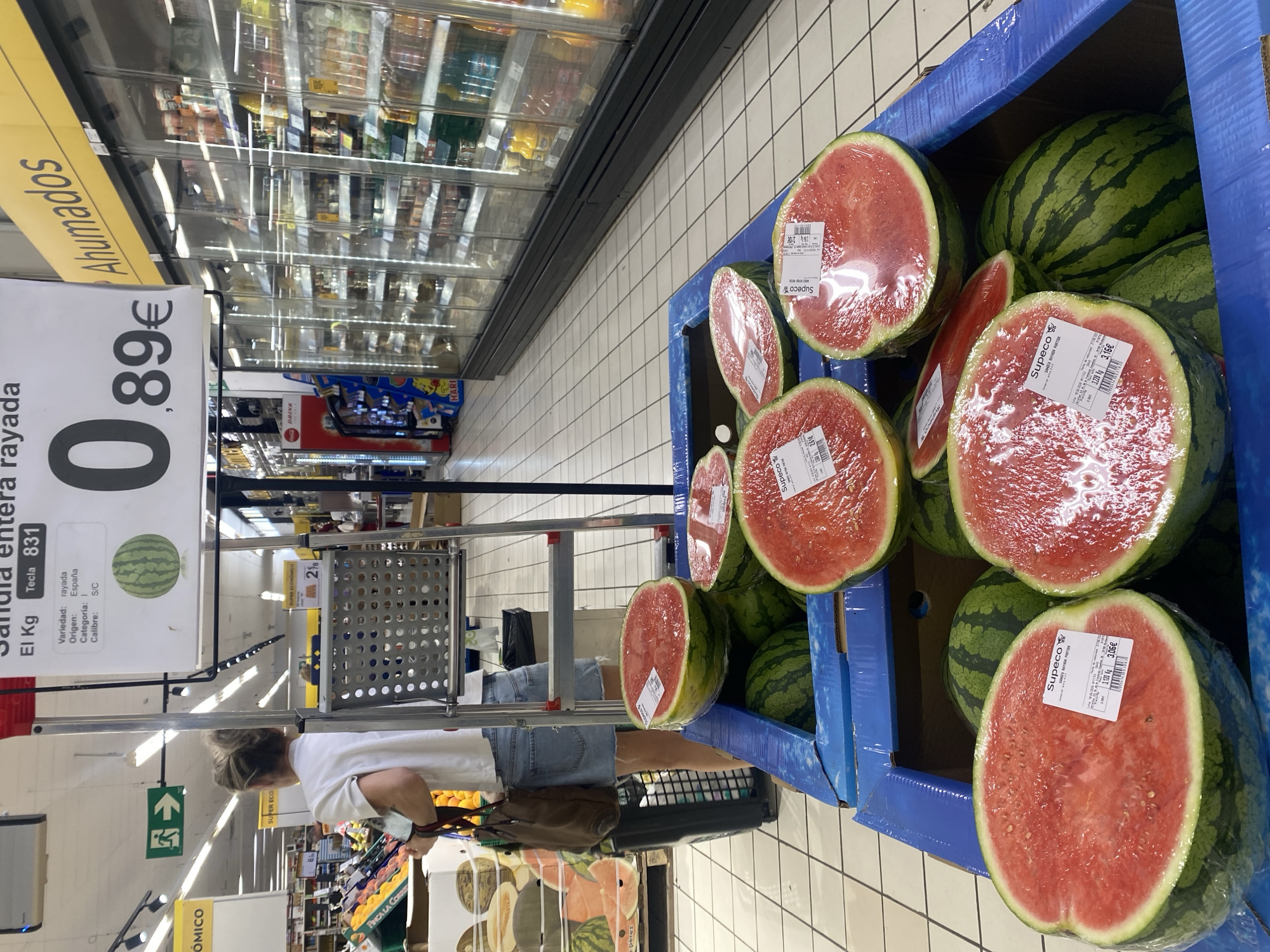 The image depicts the interior of a supermarket in Bel Air, Spain. In the foreground, several watermelons are displayed in a blue cardboard crate. Some are whole, while others are halved and wrapped in plastic. Price tags are visible on some of the watermelons. A price sign is also visible in the upper portion of the image, indicating "Sandia entera rayada" (striped whole watermelon) priced at 0.89€. In the background, a woman wearing a white t-shirt and denim shorts is seen from behind, pushing a shopping cart. Refrigerated display cases filled with various food items are visible along the right side of the image. The floor is tiled with white squares. An illuminated green emergency exit sign is visible on the left side of the background.