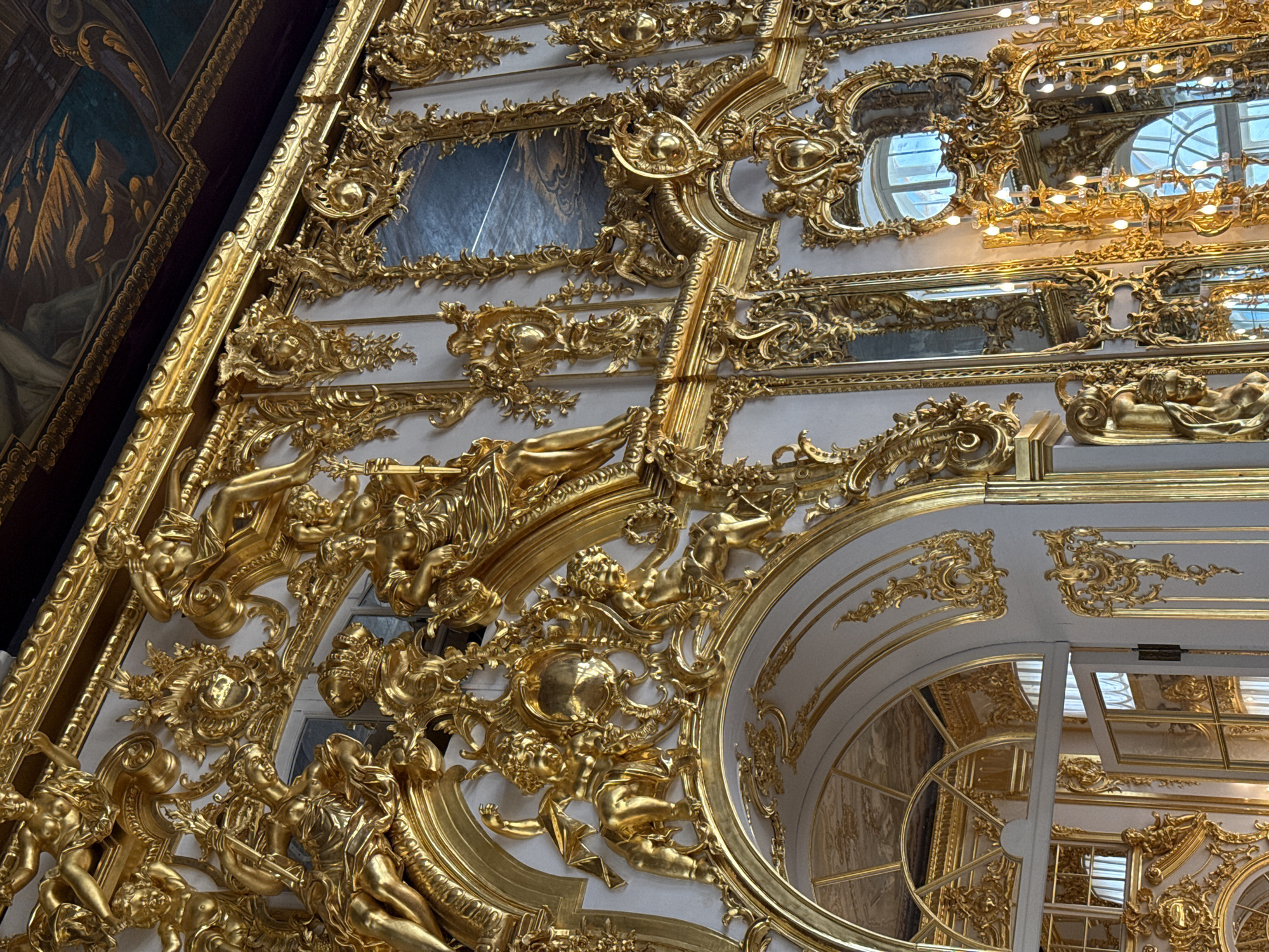 The image shows a lavishly decorated interior, likely part of the Catherine Palace in Pushkin, Russia. The walls are adorned with intricate gold ornamentation, featuring cherubs, mythological figures, floral motifs, and ornate frames. Several large mirrors are set into the walls, reflecting the opulent surroundings and architectural details. The ceiling, visible at the top of the frame, features a painted mural. The overall impression is one of extreme wealth and elaborate Baroque or Rococo design.