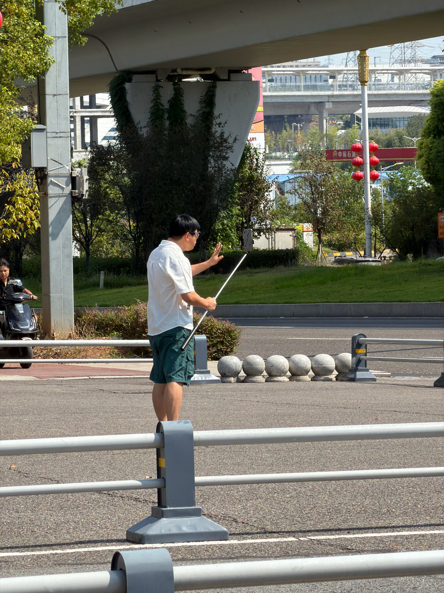 The image captures a sunny daytime scene in Jiangjin, China, likely near a road or intersection. In the foreground, a man is standing facing away from the camera, holding a white selfie stick with a phone attached. He is wearing a white short-sleeved shirt and green shorts, and he appears to be filming or taking a picture with his phone. Behind him, a woman on a black scooter is visible, also facing away from the camera. The background features a grey overpass, trees, and some buildings. Red lanterns are hanging from a pole on the right side of the image. A series of rounded stone bollards are lined up on the side of the road in front of a metal railing. The overall environment suggests an urban setting with ongoing traffic or pedestrian activity.