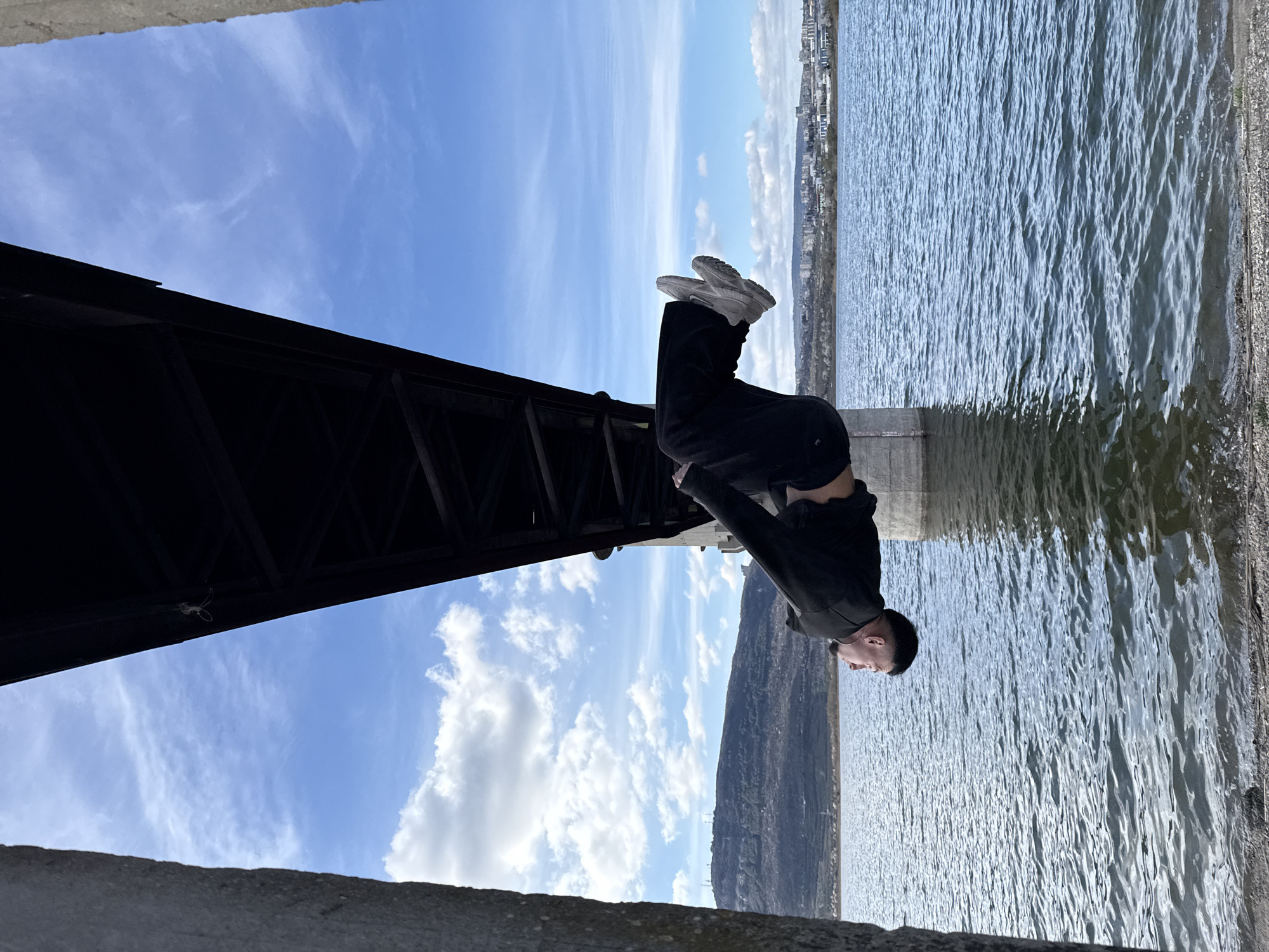 A man in a black sweatshirt and black pants is bent over backward, upside down, under a bridge. His sneakers point towards a cloudy sky. He is near a large body of water with city buildings visible in the distance on the opposite shore. The bridge is constructed of steel. The environment includes concrete structures, likely part of the bridge supports or nearby infrastructure. This photo was taken on the territory of the city of Shumen in Bulgaria near the shoreline.