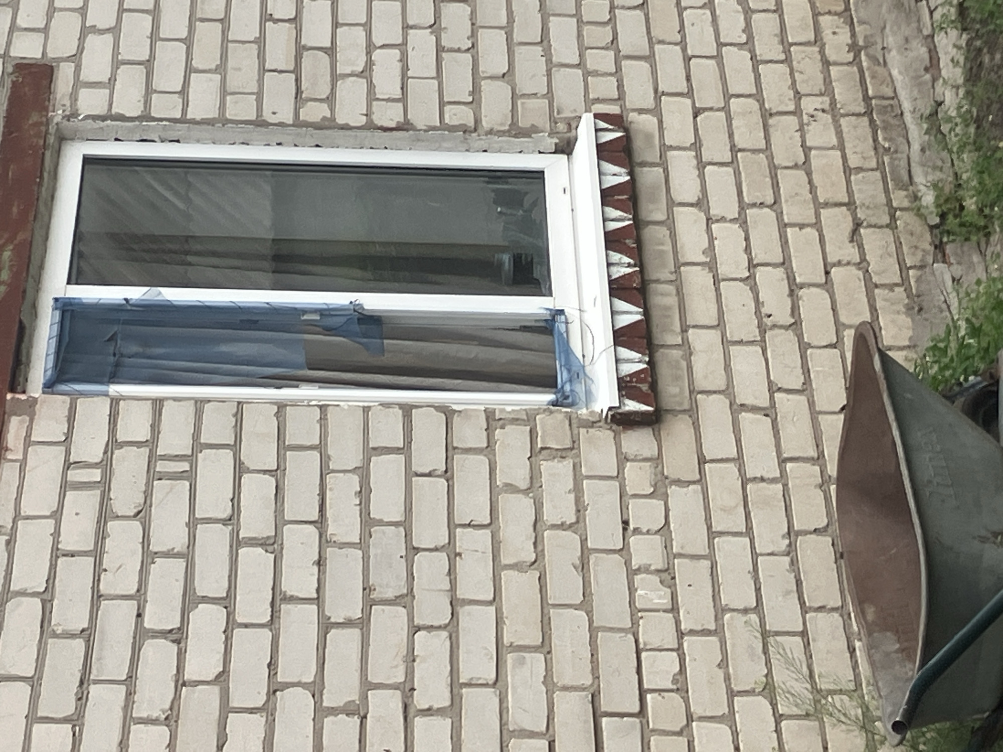 The image shows a close-up of a light-colored brick wall with a white-framed window. The window has blue netting partially covering it and grey curtains behind. Below the window is a white windowsill, and beneath that, a decorative band of alternating red and white triangular shapes is visible. A portion of a rusty brown wheelbarrow is in the foreground at the bottom left of the frame, with some green foliage to its right and in the bottom right corner. The scene is likely an exterior wall of a building.
