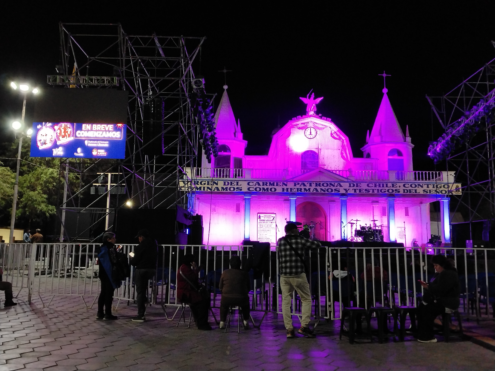 This is a nighttime photograph of the Church of Nuestra Señora del Carmen in Pozo Almonte, Chile. The church facade is illuminated with bright pink and purple lights. A banner above the church entrance reads "Virgen del Carmen Patrona de Chile, Contigo Caminamos Como Hermanos y Testigos del Señor". In front of the church, a stage and scaffolding with lights are set up. A large digital billboard displays "EN BREVE COMENZAMOS" (We'll start soon) with festive graphics. Several people are visible in the foreground, some standing and some seated on chairs behind a white metal barrier. The ground is paved with interlocking blocks. The overall atmosphere suggests an outdoor event or celebration.