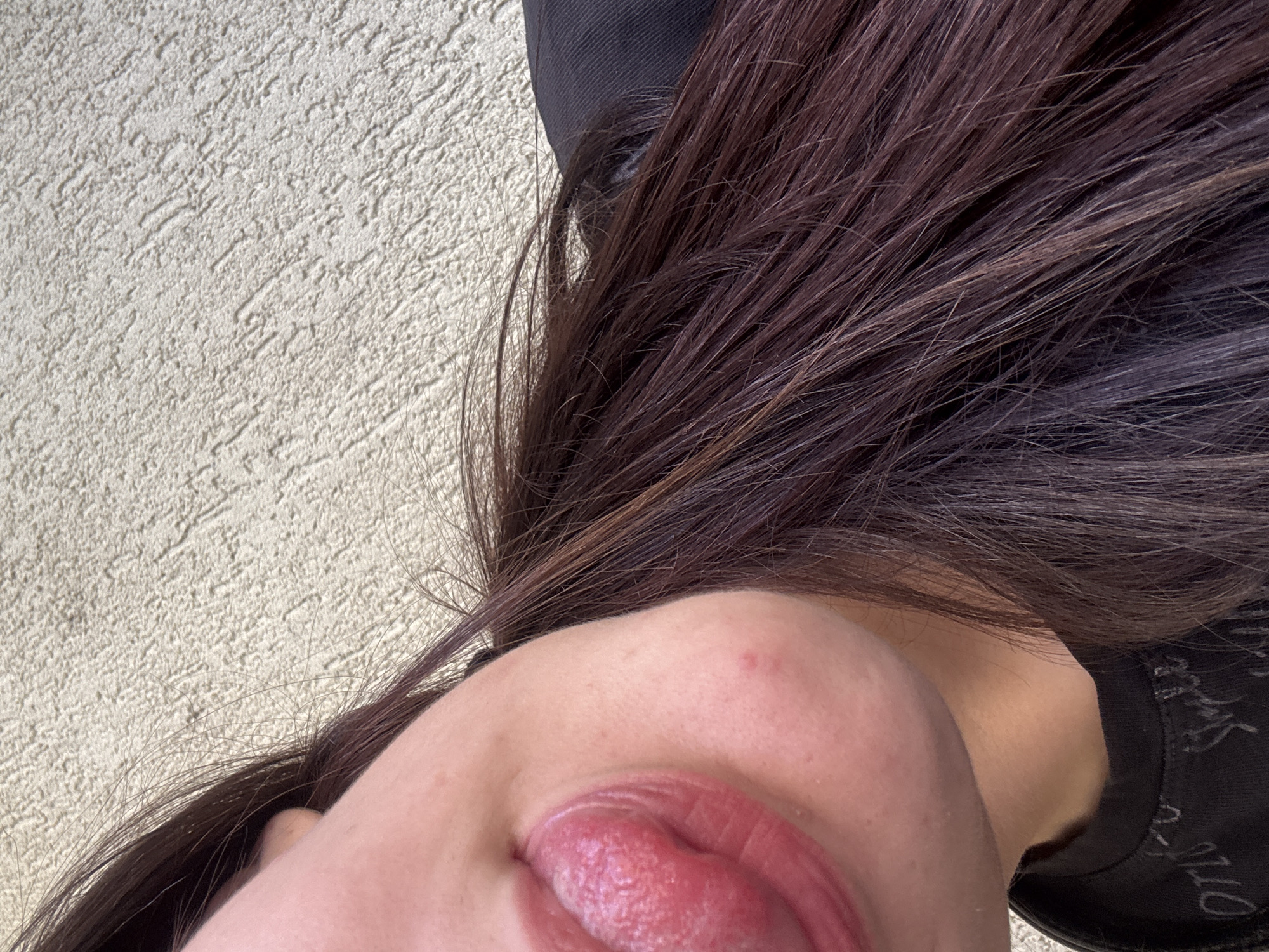 A close-up, low-angle shot captures a person's face with their tongue sticking out, against a textured wall. The individual has dark brown hair that cascades over their shoulder, obscuring part of their face. A portion of their neck and a black shirt with some writing on it is visible. There is a small pimple on the person's cheek. The background is a rough, light-colored stucco wall. The image is from Tryavna, Bulgaria.