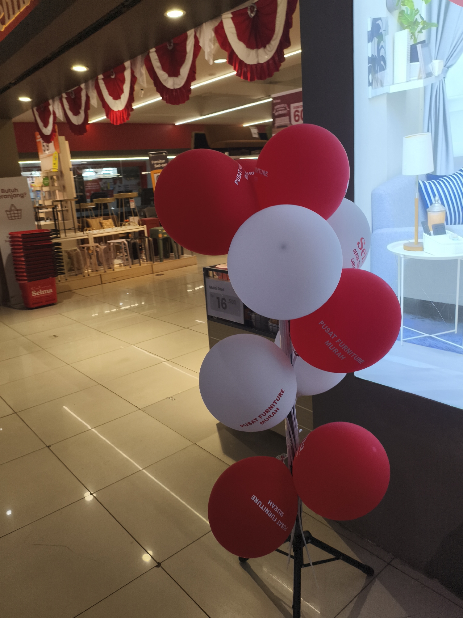 The image depicts a display of red and white balloons, some of which are printed with the text "PUSAT FURNITURE MURAH" and "SE... DEPOT... LANT..." likely indicating a furniture store in Depok, Indonesia. The balloons are arranged on a stand in the foreground. In the background, there are shelves displaying various furniture items, and a digital display showing "60" and "Mulai dari 16.500". The floor appears to be tiled and illuminated by artificial lights, reflecting on its surface. Red and white bunting is visible along the ceiling in the background, contributing to a festive atmosphere. The overall scene suggests an indoor commercial space, possibly a retail store or an exhibition hall.