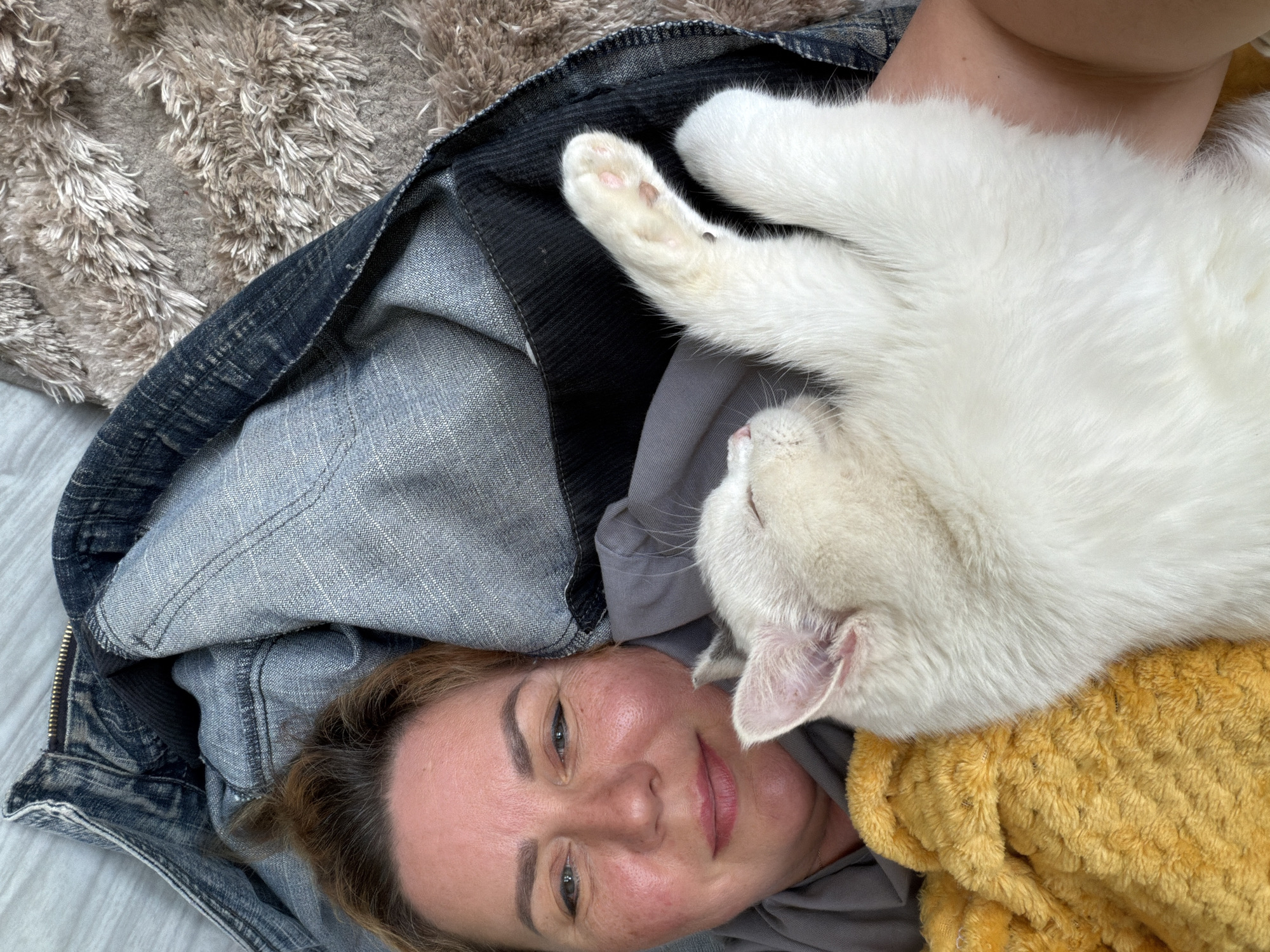 A woman lies on a light gray surface, possibly a bed or floor, with her face visible and turned slightly towards the camera. Her expression is relaxed, and her eyes are open. A white cat is draped across her chest and upper body, its paws visible in the upper left. The cat appears to be resting or sleeping. Part of a dark denim jacket or pair of pants is draped to the left of the woman and cat. A yellow, textured blanket or cloth is positioned near the woman's right shoulder and the cat's head. The background features a shag rug in neutral colors. The location is Leningradskaya, Russia.