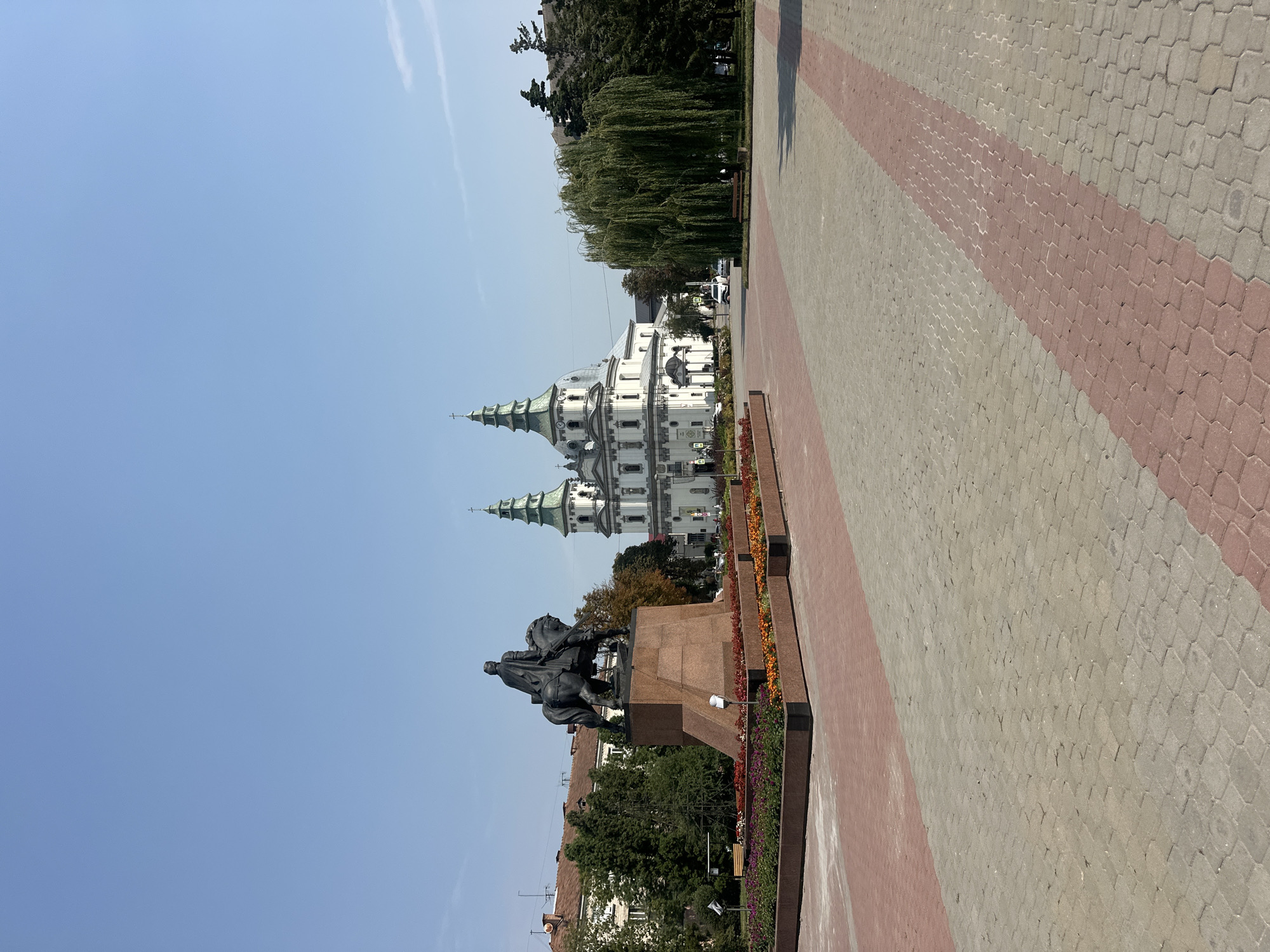 The image depicts a sunny day in Ternopil, Ukraine, featuring the Ternopil Nativity Cathedral and a monument. In the foreground, a paved plaza with a geometric pattern of light grey and red bricks leads towards a large, ornate monument topped with a bronze equestrian statue. The monument is situated in front of a planter filled with colorful flowers. Behind the monument and to the left, several trees and buildings are visible, with terracotta-tiled roofs on some structures.
Dominating the mid-ground and right of the image is the Ternopil Nativity Cathedral, a white building with a green-domed central spire and two smaller green spires. The cathedral has an elaborate facade with columns and arched windows. Adjacent to the cathedral, a weeping willow tree with long, drooping branches stands in a grassy area. Several cars are parked and driving in the background, and a wooden bench is visible near the willow tree. The sky is clear and a vibrant blue.
