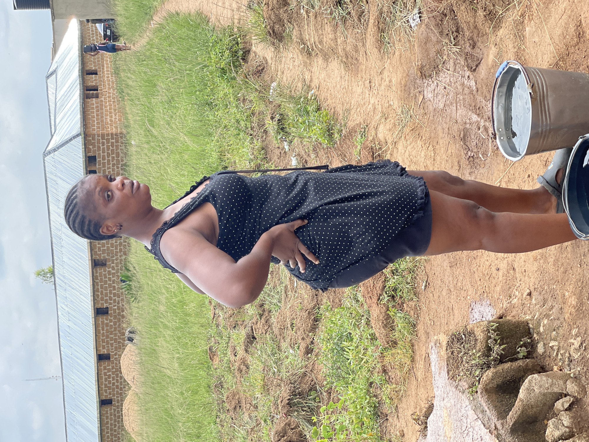 In Durumi, Nigeria, a woman with braided hair stands outdoors, facing right, her hand on her hip. She wears a black dress with white polka dots and dark shorts. In the foreground, a metal bucket filled with water sits on the ground next to her feet. A second partially visible bucket is nearby. The background features a dirt path winding through tall grass, leading towards a brick building with a corrugated metal roof. The sky is overcast. Further in the distance, another woman carries a bucket on her head, and a car is parked. The environment appears to be rural or semi-developed.
