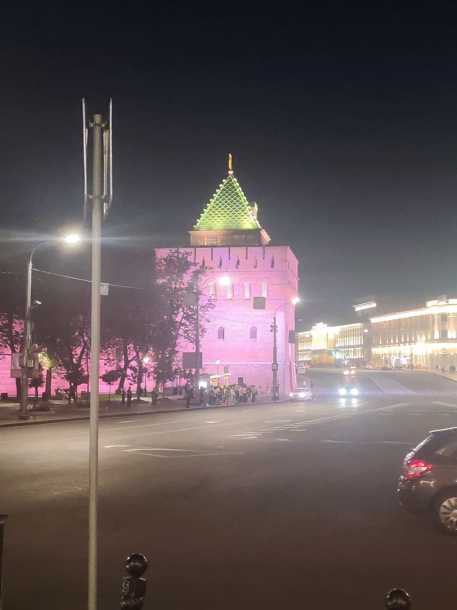 This image depicts a nighttime scene in Nizhny Novgorod, Russia, specifically near the Chkalov Stairs. On the left, a red brick tower with a green, ornate roof illuminated by pink lights dominates the frame. A tall, slender lamppost stands in the foreground, partially obscuring the tower and the trees lining a sidewalk. Several people are gathered on the sidewalk in front of the tower, illuminated by the pink light.
To the right of the tower, a wide street curves away from the viewer, with several cars visible. One car, a dark hatchback, is parked on the right side of the street in the foreground. In the distance, a row of buildings with bright white and yellow lights lines the street, suggesting a commercial or public area. The sky is dark, indicating it is nighttime. The overall impression is of a lively urban setting at night, with historical architecture illuminated for effect.