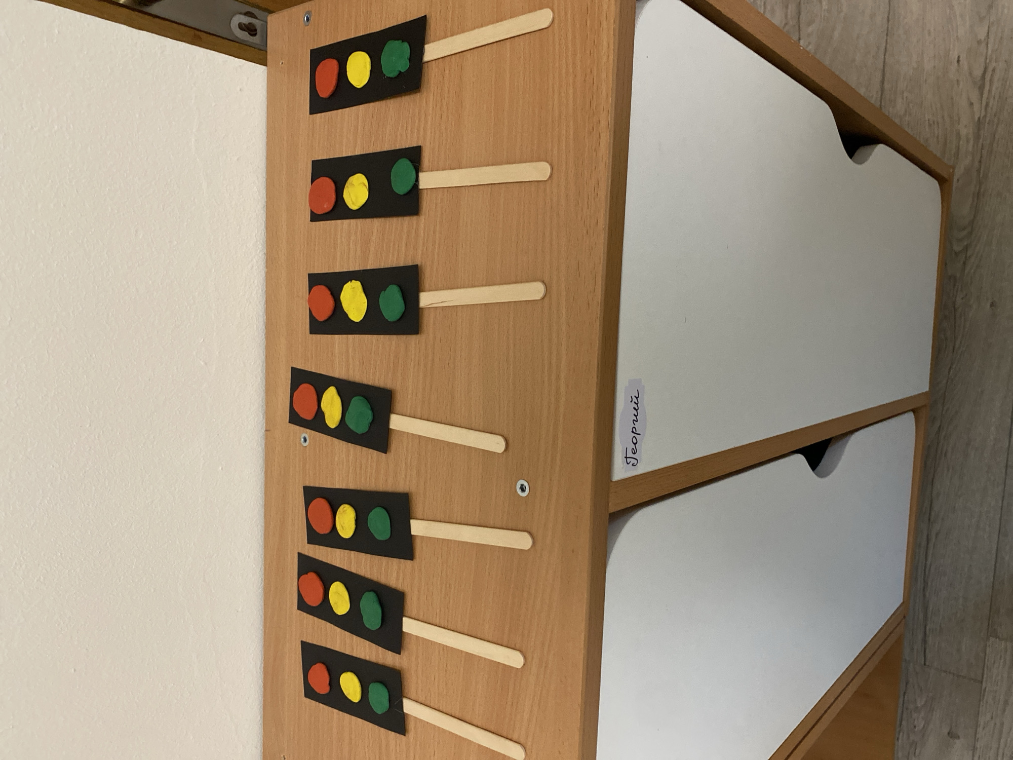 The image shows a wooden cabinet with white drawers. On the side of the cabinet are multiple traffic light depictions, each consisting of a black rectangle with three colored circles (red, yellow, green) arranged vertically. Each traffic light depiction is attached to a wooden stick. The side of the cabinet is inscribed with the name "Георгий" (Georgiy in English), written vertically. The scene is indoors, and based on the name inscription and available information, it can be inferred that the location is likely in Sochi, Russia.