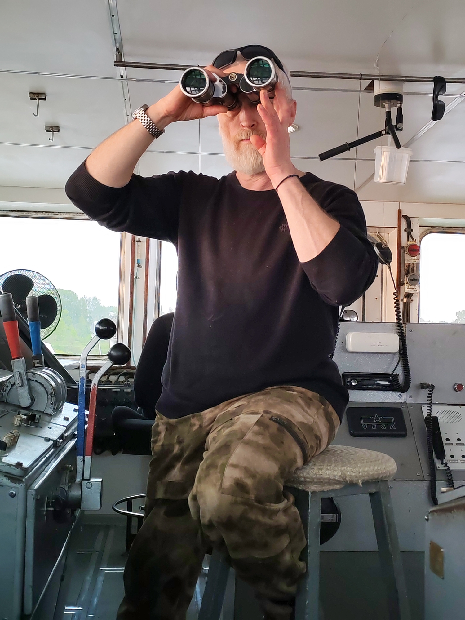 A man is seated on a stool inside a cabin, possibly a boat's wheelhouse. He has a gray beard and is wearing a black long-sleeved shirt and camouflage pants. He is holding binoculars up to his eyes, looking out. He also has sunglasses perched on his head and is wearing a watch on his left wrist. The cabin contains control panels, levers, and a fan. Outside the window, there is a glimpse of a river landscape with trees and greenery. Based on the image description, the location is Нифантово, Russia.