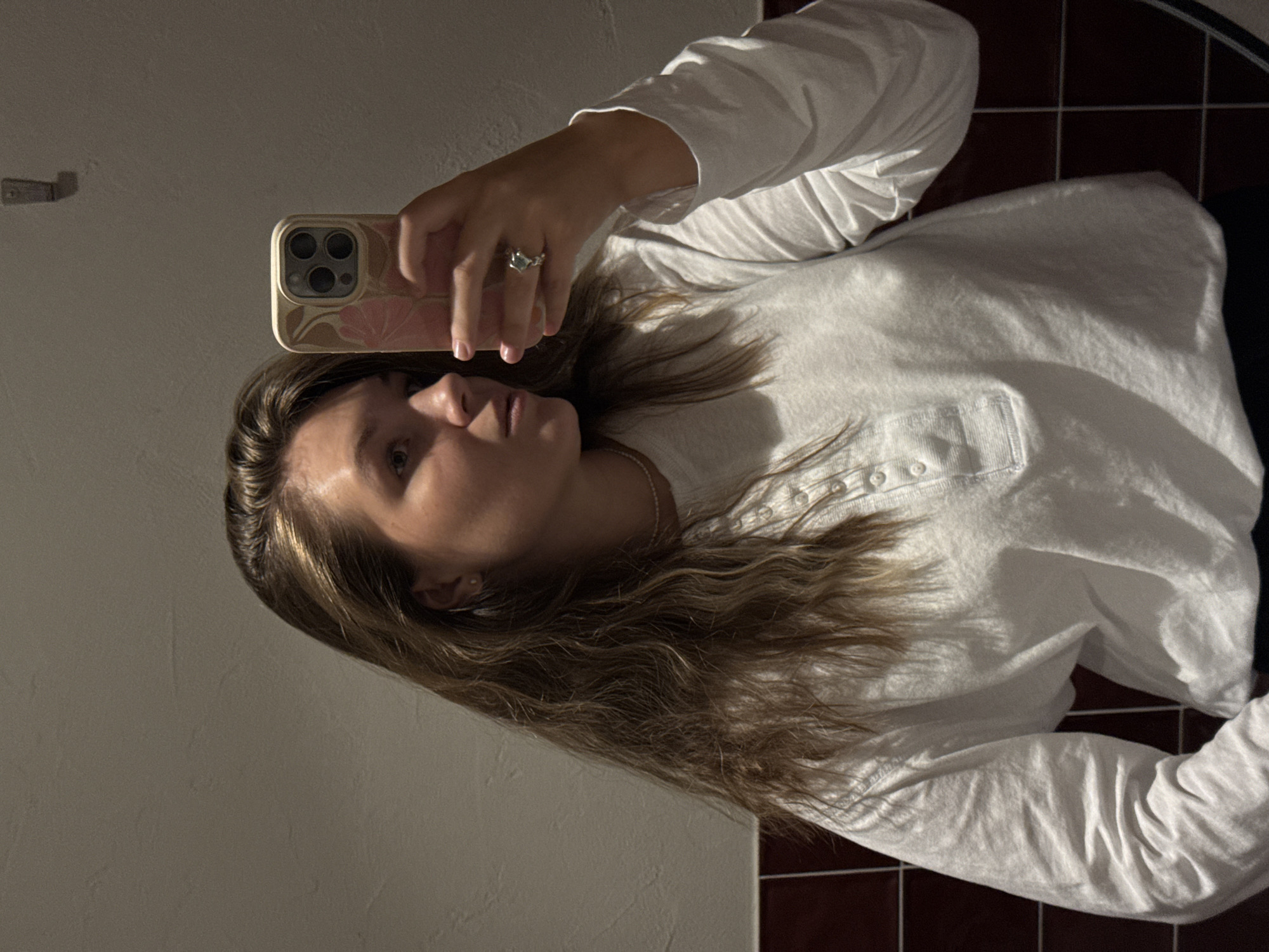 The image shows a young woman with long, light brown hair taking a selfie in a bathroom. She is wearing a long-sleeved white henley shirt. She has a ring on her right hand, which is holding a smartphone. The bathroom walls are tiled with light beige tiles and a darker red border near the bottom. The lighting is dim, casting shadows. There is no indication of the city of Belgrade, Serbia, in the image.