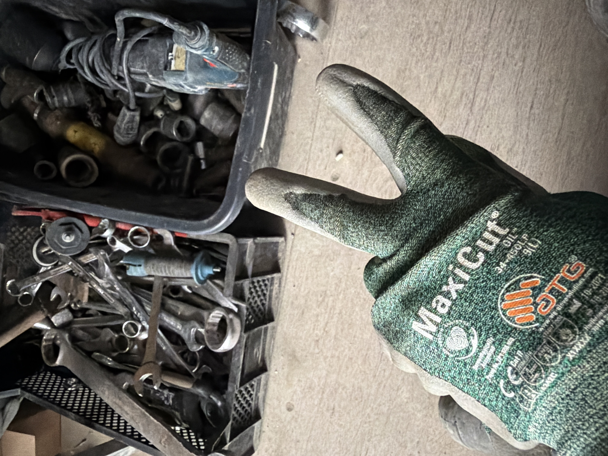 A gloved hand makes a peace sign in the foreground, with two fingers extended. The glove is green with gray accents and bears the text "MaxiCut" and "OTG". The background shows two black plastic bins filled with tools, including wrenches and sockets. The scene appears to be in a workshop or garage. The image is an outdoor shot, with a wooden floor visible beneath the bins and hand. The lighting suggests it is daytime. There are no visible signs of the city of Janowice, Poland.