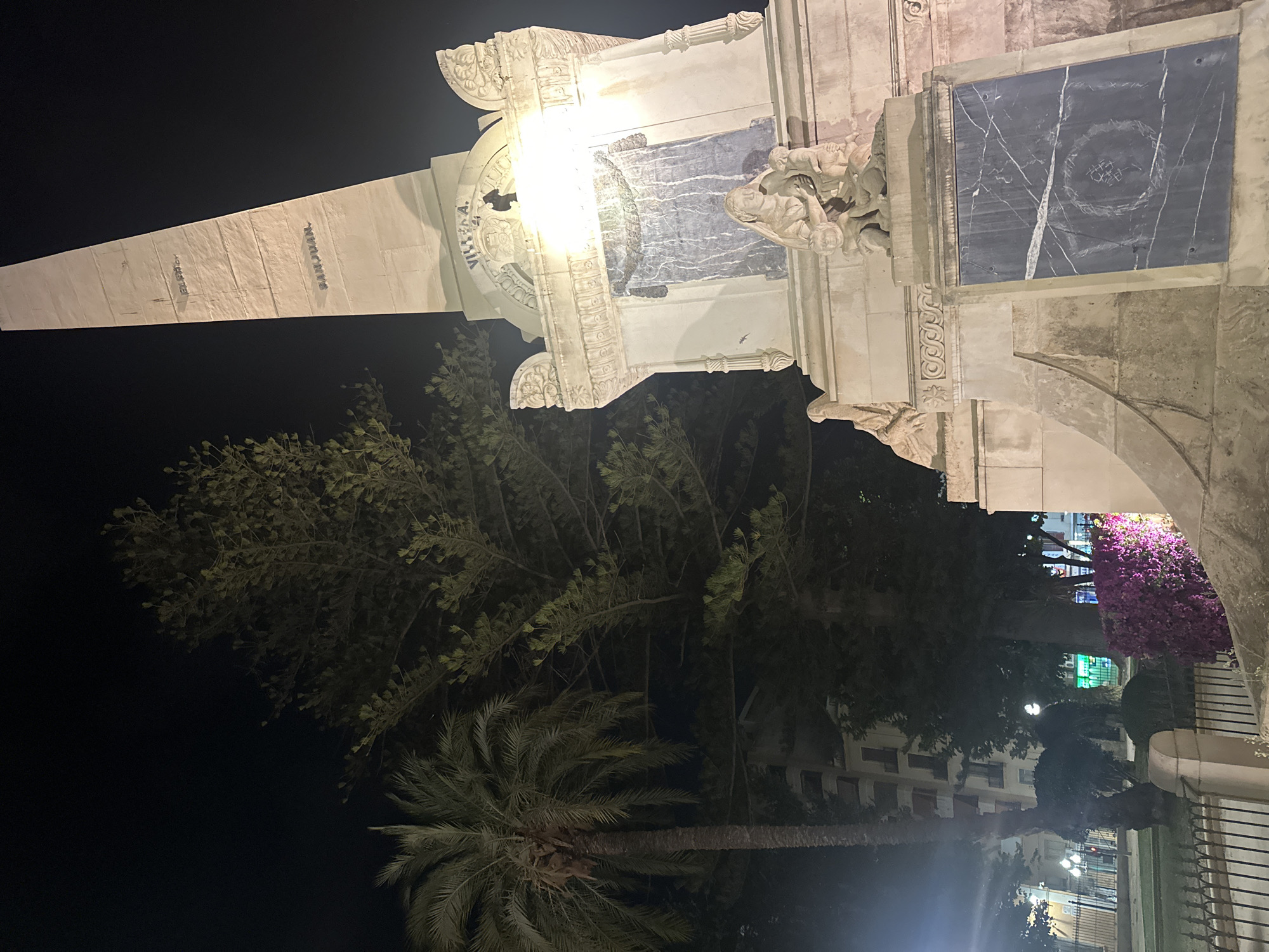 This image shows a monument at night in Alicante, Spain. The monument is made of stone and has inscriptions. There are also sculptures on the monument. In the background, there are trees, buildings, and streetlights. The monument is illuminated by artificial lights. The overall scene is dark, with highlights from the lights.