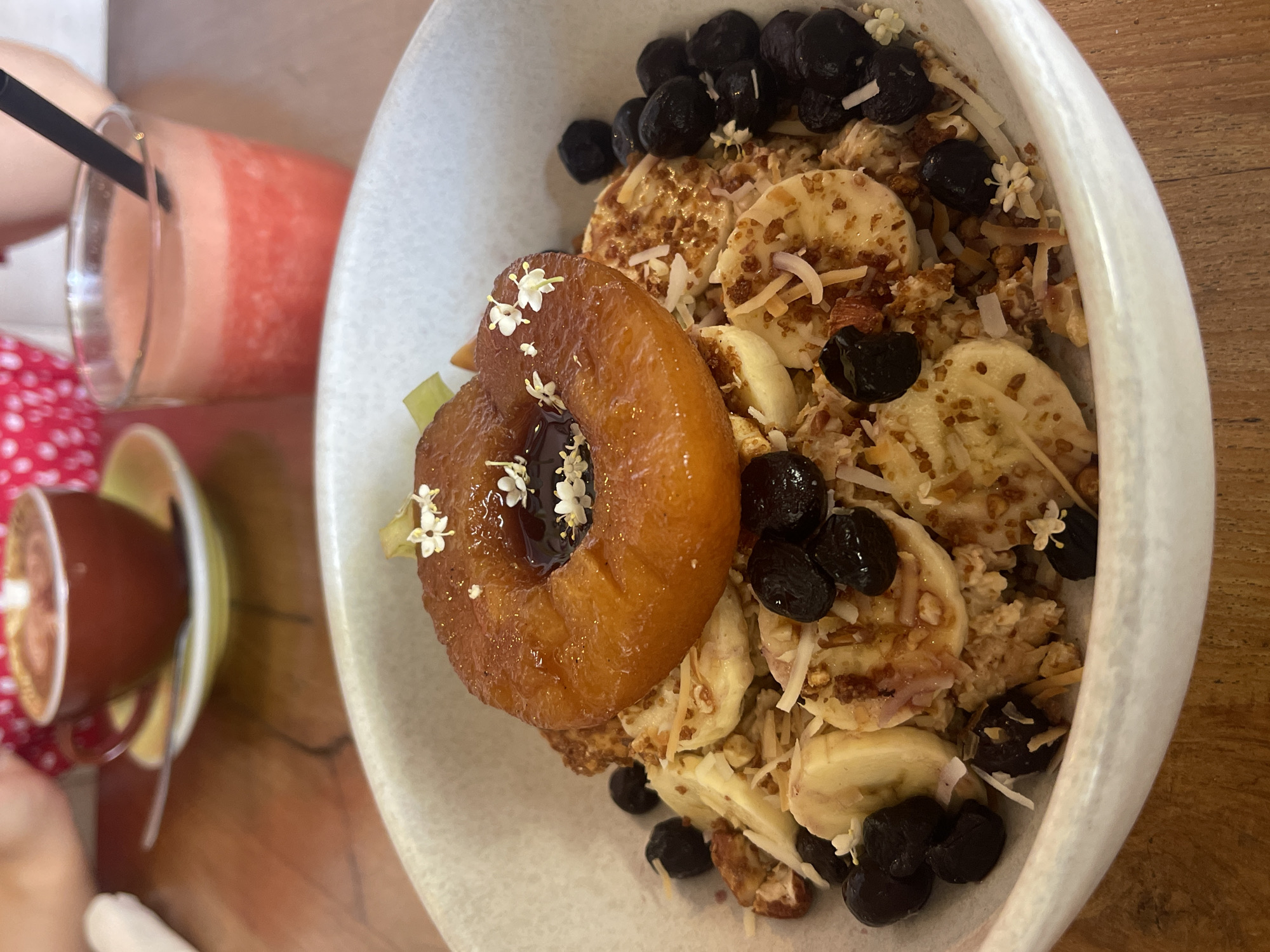 A medium-shot captures a breakfast setting, featuring a bowl of granola, banana slices, blueberries, and a torched piece of fruit topped with small white flowers, all within a white bowl. A small cup of coffee and a pink smoothie with a black straw sit alongside the bowl on a wooden table. The photograph was taken in North Kuta, Indonesia.