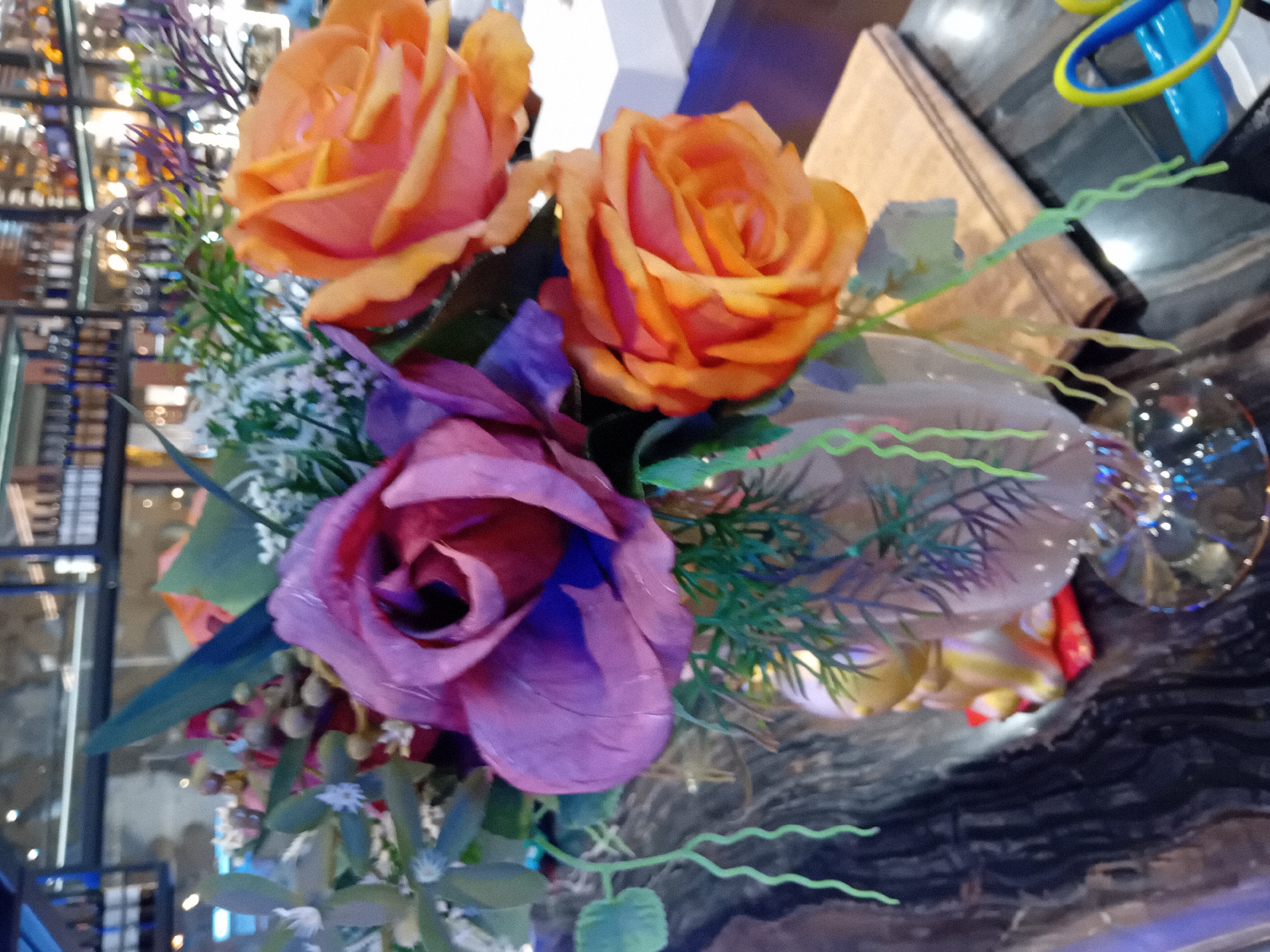 The image presents a close-up of a vibrant artificial floral arrangement, prominently featuring three large roses. Two roses are a warm blend of orange and peach hues, while a third, slightly larger rose displays shades of purple and magenta. These central flowers are surrounded by an assortment of artificial greenery, including delicate light green wavy strands, darker green fern-like foliage, and small clusters of white and purplish filler flowers.
The background is softly blurred, indicating a shallow depth of field, and suggests an indoor setting. On the left side, indistinct, illuminated shelves with various objects can be seen, possibly hinting at a retail space or restaurant. To the right, a light brown object resembling a book or a small wooden box is visible, along with reflective surfaces and a portion of a clear glass item, such as a wine glass. In the upper right corner, bright blue and yellow plastic rings or decorative shapes are partially discernible. The overall impression is a decorative display, emphasizing the colorful artificial blooms against a softly lit, busy interior backdrop. There is no visible text.