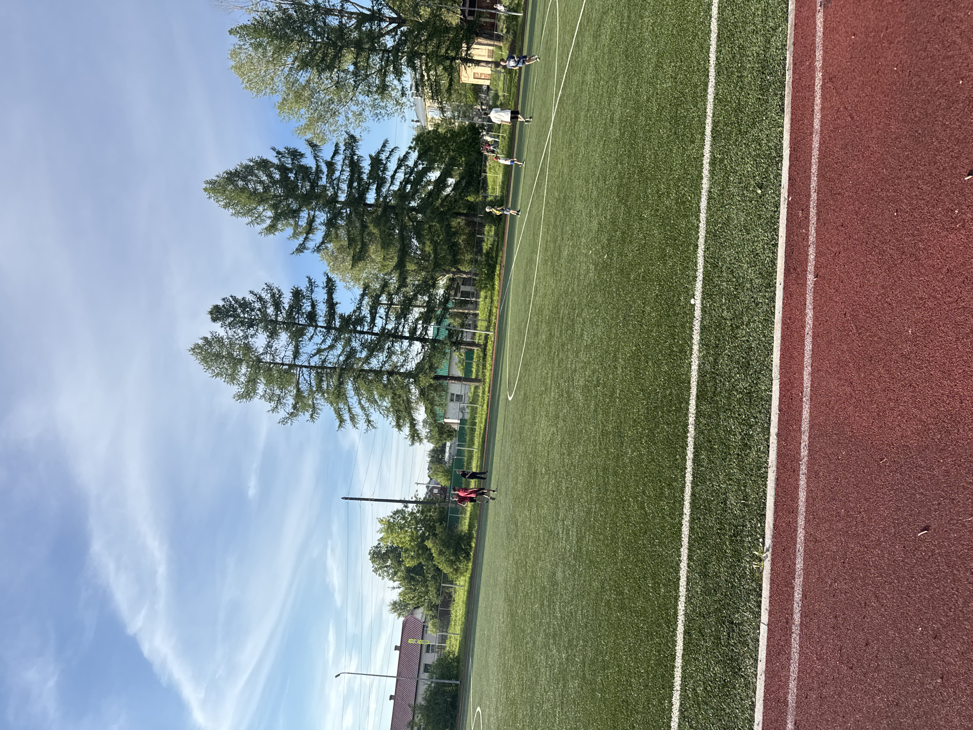 The image depicts an outdoor artificial turf soccer field in the city of Asha, Russia. A group of people, appearing to be children and young adults, are engaged in a soccer-related activity on the field. Some are in motion, suggesting they are playing or practicing. In the background, residential buildings, trees, and power lines are visible, indicating an urban or semi-urban environment. The sky is clear with some wispy clouds. A red running track borders the soccer field in the foreground.
