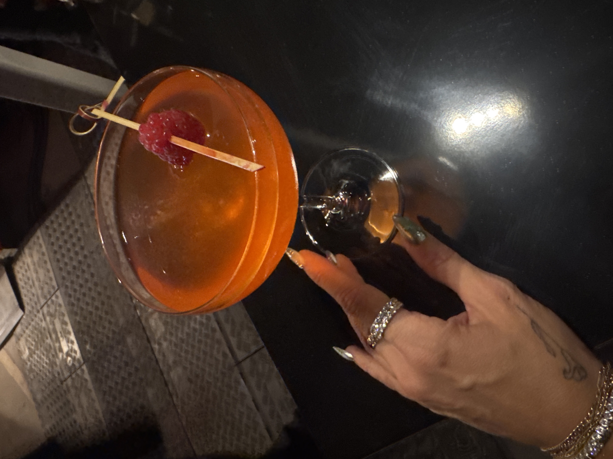 The scene depicts an overhead shot of a cocktail in a stemmed coupe glass. The drink has an orange hue and is garnished with a raspberry speared on a toothpick. A hand, adorned with a diamond ring and bracelets, reaches for the glass. The location appears to be a dimly lit indoor setting with a dark surface, possibly a table or bar. The presence of the cocktail and jewelry suggests a social setting, possibly a bar or restaurant. Given the information and limitations of the image, it is not possible to pinpoint the location in Indio, United States of America.