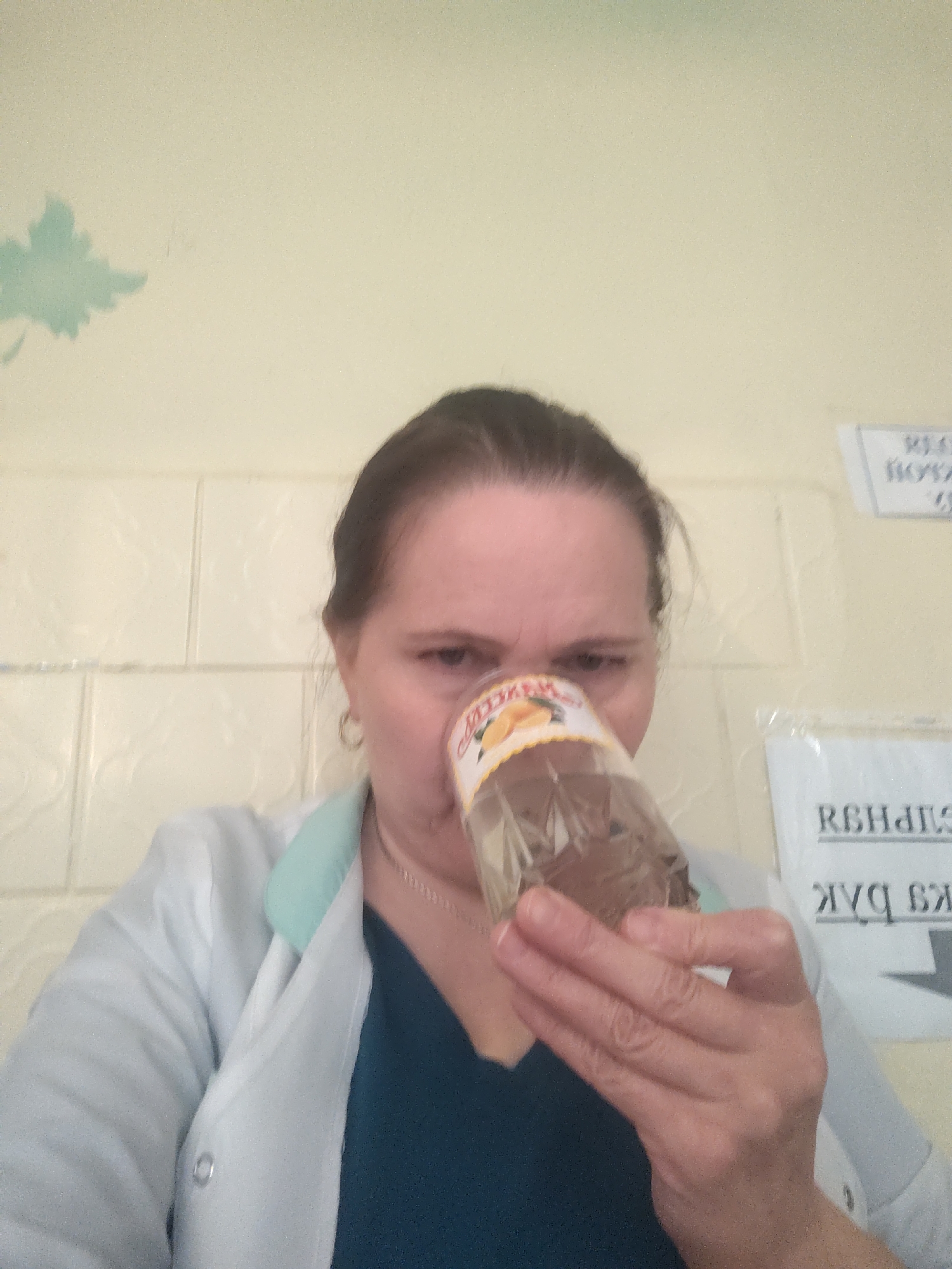 The image depicts a woman, likely middle-aged, indoors in what appears to be an institutional or medical setting in Sarapul, Russia. She has brown hair pulled back and is wearing a white lab coat with a light blue collar over a dark teal shirt, complemented by a silver chain and a small earring.
The woman is actively smelling or inspecting the contents of a translucent plastic bottle, which she holds close to her nose. Her facial expression, characterized by a furrowed brow, suggests concentration, curiosity, or possibly a reaction to the bottle's contents. The bottle's label features an orange graphic and partially visible Cyrillic text, including "НЕРЕ".
The background consists of a light-colored, possibly cream or light yellow, wall. Part of this wall is covered with textured, square tiles. A light green, leaf-shaped stencil or decal is visible on the upper left side of the wall. To the right, several white papers with Cyrillic text are taped to the wall, some appearing inverted or partially obscured, with visible fragments such as "REHBEПЧ" and "Я БАК". The scene is illuminated by indoor lighting.