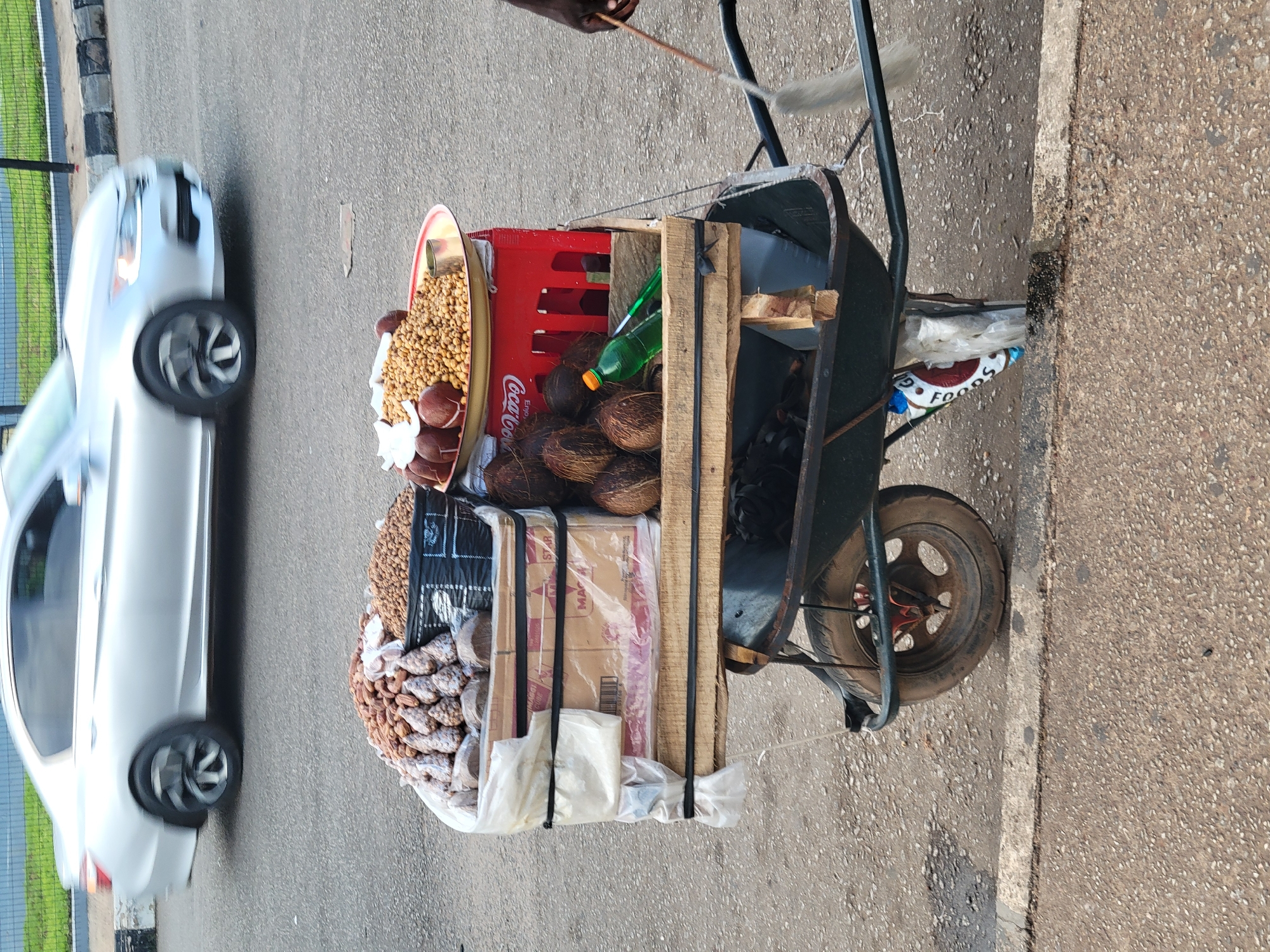 An outdoor scene in Barakin Lerawi, Nigeria, features a street vendor's cart. The cart, a modified wheelbarrow, holds various goods for sale. In the foreground, the cart's dark green basin is visible, with a white plastic bag secured to the front wheel and a portion of a red and white branded bag partially visible. Resting on the cart's wooden platform are several coconuts, a green plastic bottle with an orange cap, and a red Coca-Cola crate. On top of the crate is a shallow, circular tray containing a pile of small, light brown nuts and some reddish-brown round fruits, possibly dates, with a small metal cup. Behind this, two black plastic containers are filled with different types of nuts. One container holds numerous small, dark brown nuts, while the other holds a mix of larger, lighter brown nuts and what appear to be dried fruits resembling dates. A silver sedan is in motion in the background, partially obscuring the road and a striped curb. The ground is paved with asphalt.