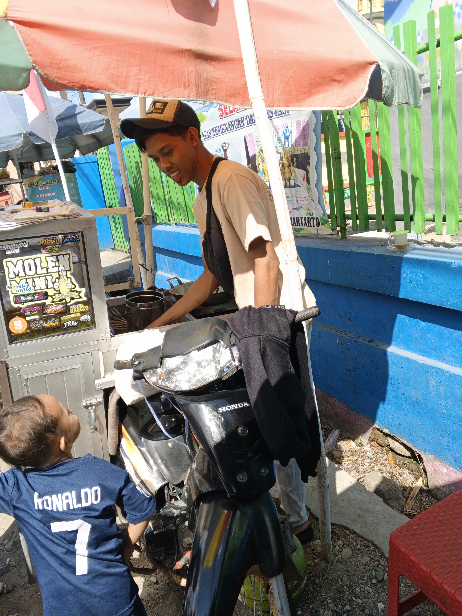 This is a medium shot, taken from a low angle, of a young boy wearing a blue jersey with "RONALDO 7" printed on the back, looking up at a vendor. The vendor, a young man wearing a tan t-shirt and a black baseball cap, is standing behind a food cart and appears to be preparing food. He is smiling and looking down at the boy. The food cart is a custom-built mobile unit on a motorcycle chassis, with a bright orange and green umbrella providing shade. The cart has various stickers on its side, advertising "Molen Mini" and other products. In the background, there is a bright blue wall and a green picket fence, suggesting an urban environment, possibly in the city of Padang, Indonesia, known for its street food culture and vibrant colors. The ground is covered in gravel and dry leaves. A red plastic stool is visible in the lower right corner.