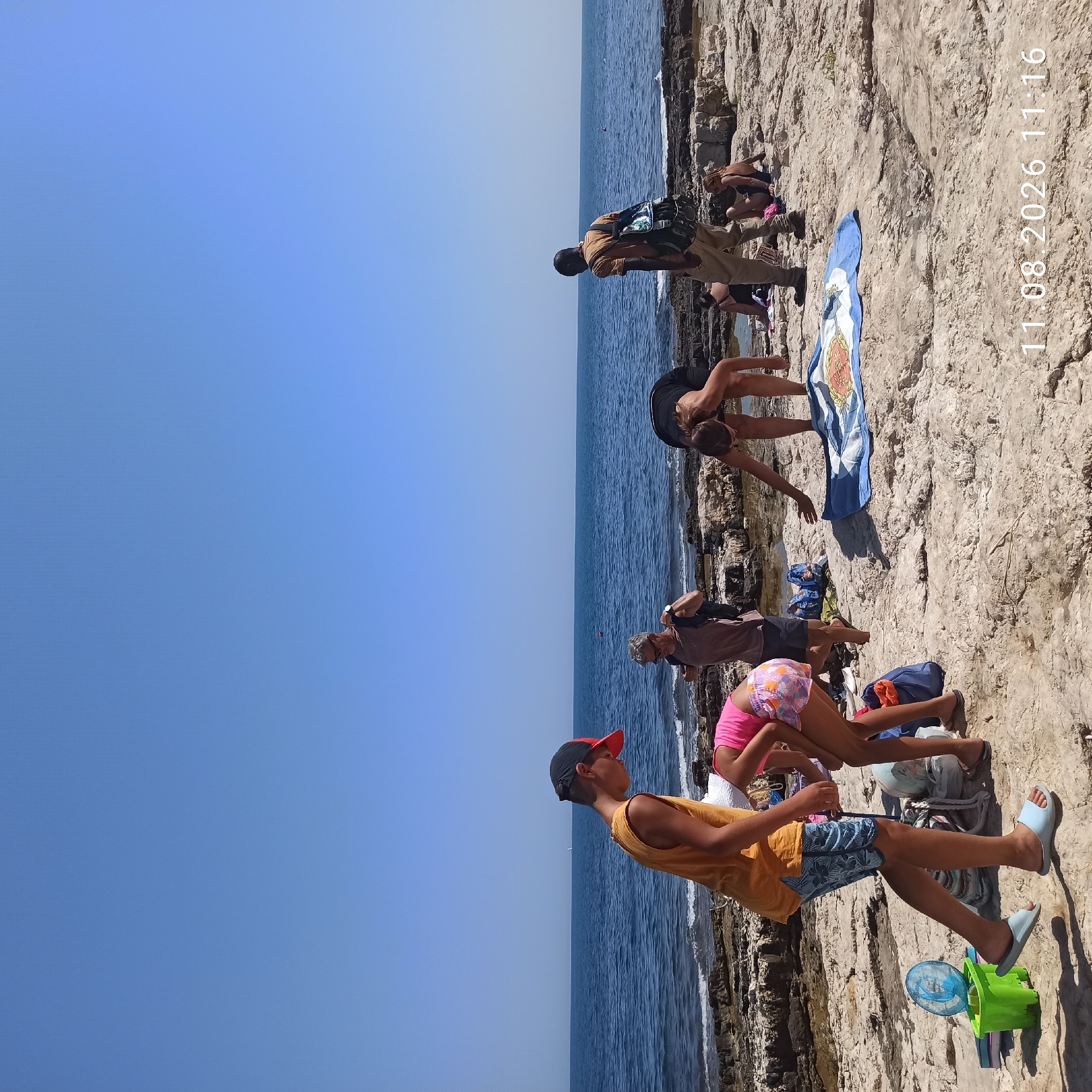 On a sunny day, a group of people are gathered on a rocky shoreline in Molfetta, Italy, with the Adriatic Sea in the background. A young boy in a yellow tank top and blue patterned shorts, wearing a red and black baseball cap and blue flip-flops, walks in the foreground. To his right, a young girl in a pink tank top and colorful patterned shorts is bent over. Further back, a man with gray hair wearing sunglasses and a brown shirt with a backpack is standing. To his right, a woman with dark hair is bending over, and behind her, a person with a backpack is walking away. In the distance, several people are seated on towels on the rocks. The environment consists of light-colored rocks and clear blue water under a bright blue sky. A green bucket with a blue toy fan is near the young boy. A blue towel with an orange design is laid out on the rocks. The timestamp "11.08.2026 11:16" is visible in the lower right corner of the image.