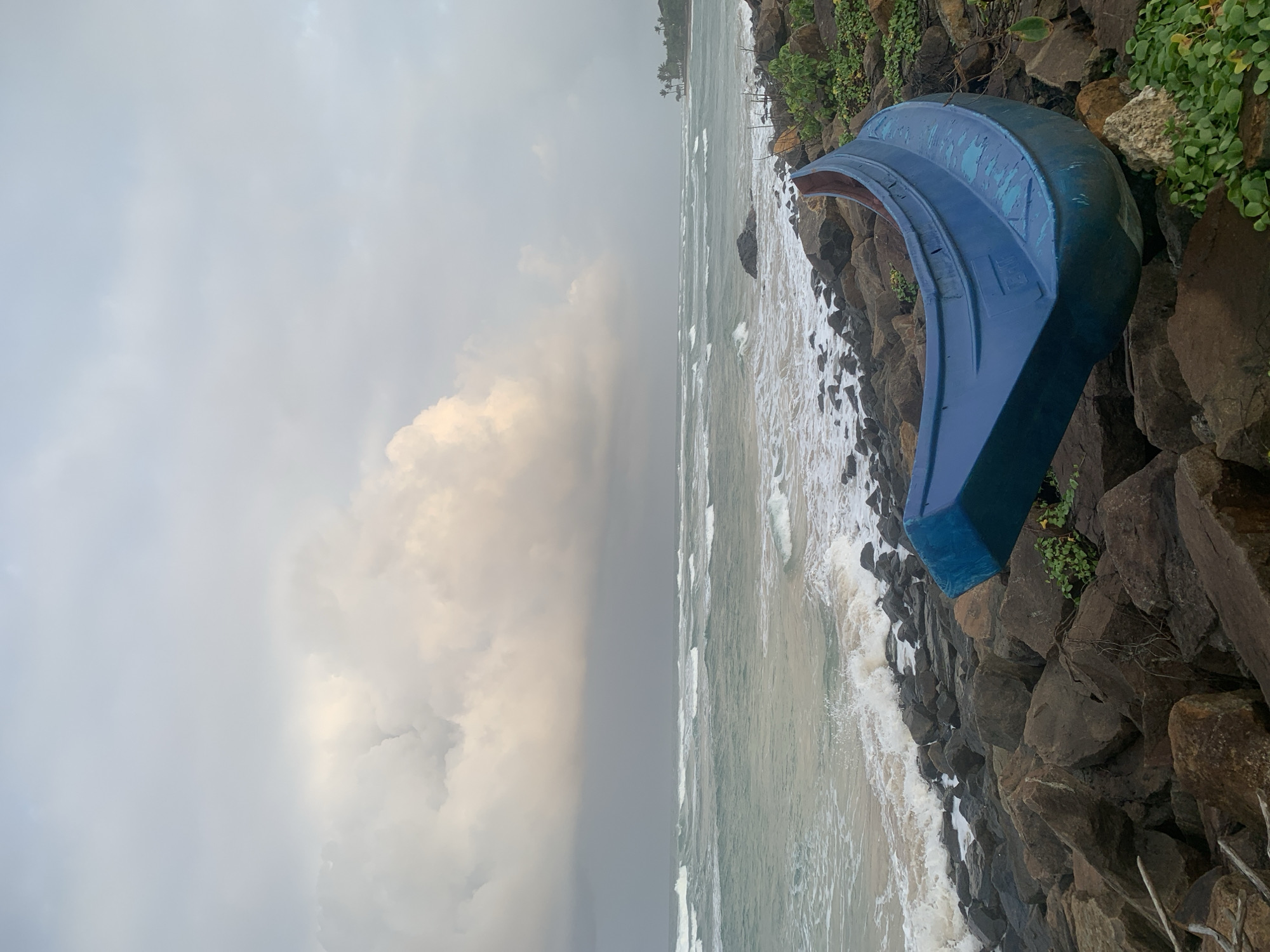 The image captures a serene coastal scene in Kathaluwa, Sri Lanka, under a cloudy sky. In the foreground, a weathered, bright blue boat rests on a rocky shore, its hull showing signs of age. The choppy sea stretches out to the horizon, with waves crashing against the rocks. Lush green vegetation grows among the stones, adding a touch of life to the rugged landscape. In the distance, palm trees dot the coastline, hinting at the tropical environment. The overall mood is tranquil, with the natural elements taking center stage.