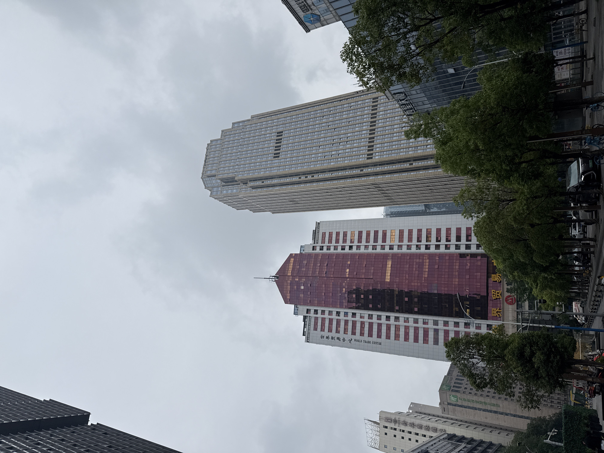 The image presents a dense urban cityscape featuring multiple tall buildings under an overcast, gray sky. In the foreground and center, a prominent skyscraper with a distinctive reddish-maroon glass facade stands out. On this building, the white text "锦绣商贸中心 WORLD TRADE CENTRE" is clearly visible, along with a partial yellow sign that reads "嘉酒店" (Jia Hotel) further down. To the left and slightly behind, another very tall building rises, characterized by its lighter-colored, multi-tiered, and somewhat blocky architectural design, featuring numerous windows.
A street or thoroughfare is visible on the right side, bordered by a line of green trees. Several parked cars and bicycles or scooters can be seen along this street. Peeking through the trees, another building displays the green text "假日酒店" (Holiday Inn). Other buildings, including one with a dark, patterned facade and another with a blue star-like logo and unreadable Chinese text, are visible in various parts of the frame, contributing to the urban density. The overall impression is of a bustling city center on an overcast day, with no specific human activity discernible beyond the implied presence in a commercial district. The location cues point to a commercial hub in a Chinese city.