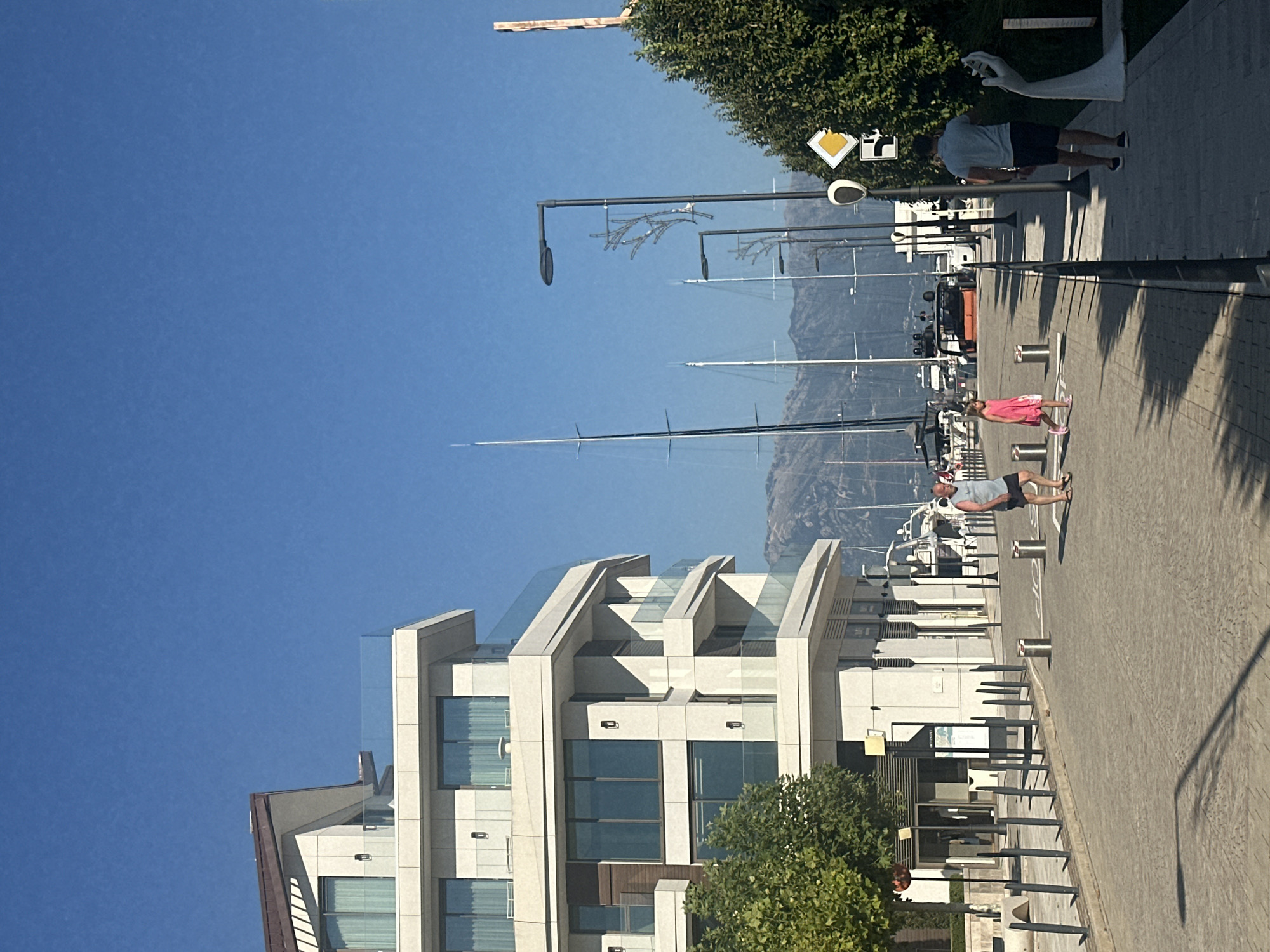The image depicts a sunny day in Kumbor, Montenegro, featuring a modern waterfront promenade with a large building on the left, likely an apartment complex or hotel, characterized by its white facade, glass balconies, and a brown roof. Several sailboats with tall masts are docked in the marina visible in the background, against a backdrop of hazy mountains under a clear blue sky. The paved promenade has bollards and streetlights. In the foreground, a man in a grey tank top and black shorts walks to the right, and a young girl in a pink dress walks to the left, followed by a man in a blue polo shirt and black shorts. A modern white sculpture of a hand is visible on the right side of the promenade. Trees line the left side of the promenade, and vegetation is present on the right.