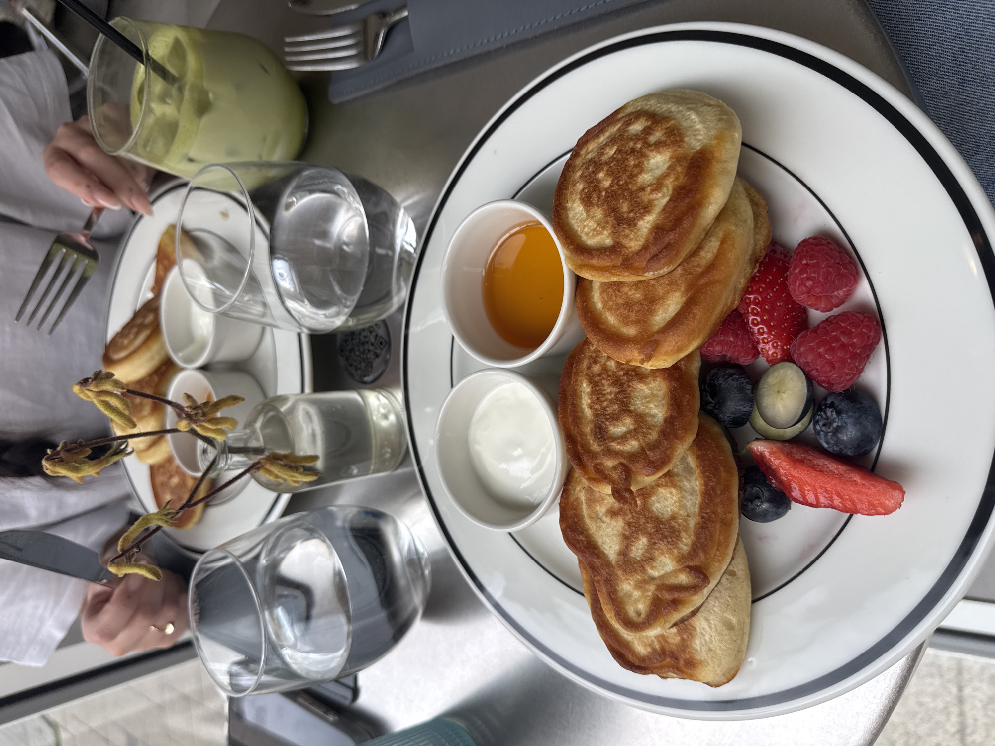 The image shows a brunch scene in a restaurant, possibly in Sofia, Bulgaria, based on observed architectural styles. Two white plates with black rims are visible. On the right plate, a stack of small pancakes is accompanied by small bowls of honey and yogurt, along with a mix of fresh berries including strawberries, raspberries, and blueberries. On the left plate, there are also pancakes with honey and yogurt. There are two glasses of water, and a green smoothie in a glass with a straw. Silverware is also present. A small vase with flowers is placed between the plates. The environment appears to be an interior setting, likely a restaurant or cafe, with a grey tabletop. Parts of people are visible at the edge of the image, with hands holding utensils. The overall scene suggests a breakfast or brunch meal being enjoyed in a modern setting.