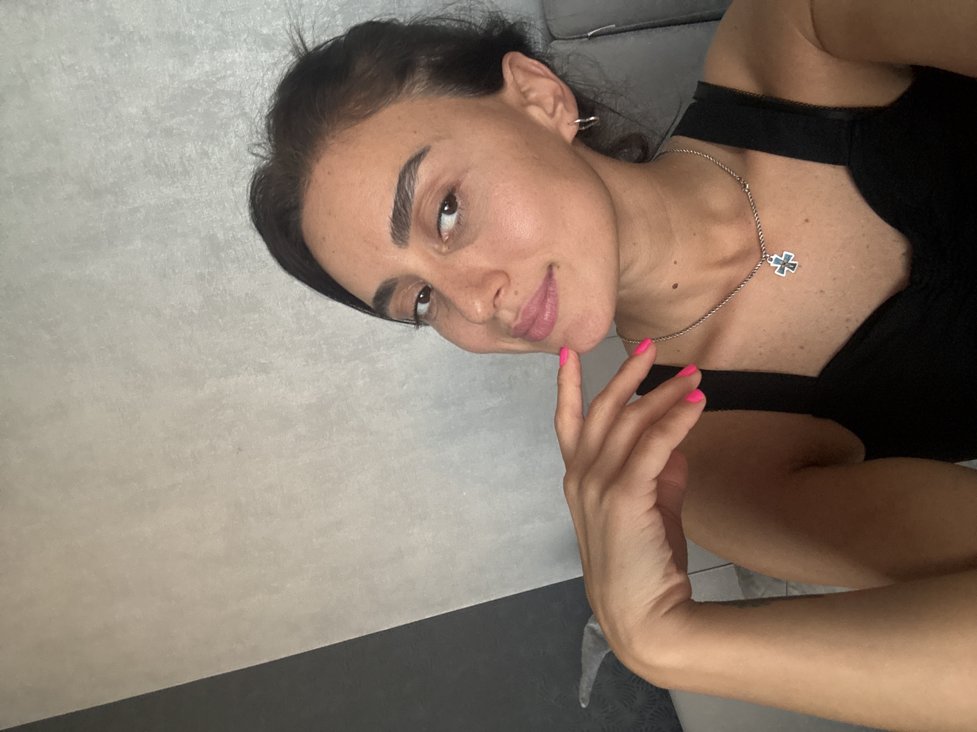 A young woman with dark hair and brown eyes is captured in a close-up, head-and-shoulders shot, looking towards the viewer with a slight smile. Her left hand, with bright pink fingernails, is gently resting on her chin. She is wearing a black top and a silver necklace with a cross pendant. The background is a textured wall, divided diagonally into a dark gray section on the left and a lighter gray section on the right. The lighting is soft, casting subtle shadows on her face. The image appears to be taken indoors, in a residential setting, possibly an apartment in Tbilisi, Georgia.