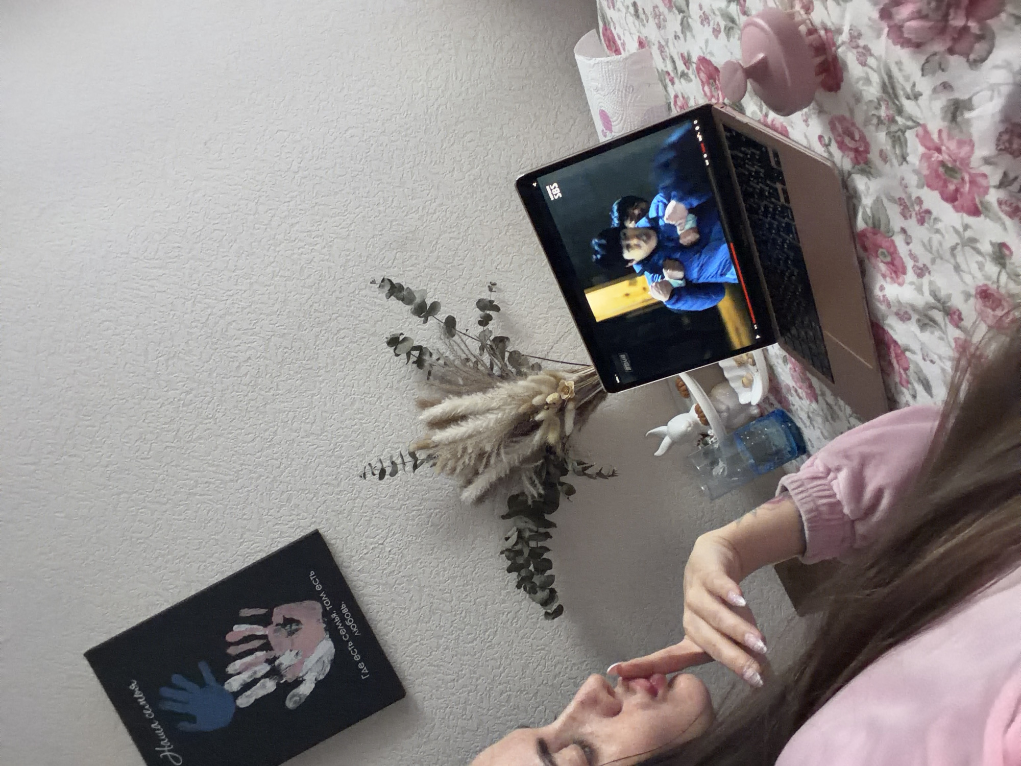 The image captures an indoor, cozy scene likely in a bedroom or living area within Saint Petersburg, Russia. A woman with long, brown hair, wearing a pink top, is depicted lying down. Her right hand, featuring nails with glittery polish, is gently placed near her mouth, suggesting a moment of relaxation or contemplation.
In the upper right, a metallic-colored laptop, possibly a MacBook Air, rests on a bed or surface covered by a pink floral-patterned bedspread. The laptop screen displays a video, identifiable by the "SBS" logo (a South Korean broadcasting network), showing two people, one in a blue puffer jacket. Near the laptop, a pink ceramic stand and a roll of white paper towels are visible.
To the left of the laptop, a dried floral bouquet, composed of eucalyptus branches and various feathery grasses, is artfully arranged against a textured, light gray wall. Below the bouquet and to the left, a black canvas or board is displayed. This board features painted handprints in blue, pink, and white, alongside prominent Russian text. The text reads: "Наша семья" ("Our family") and "Где есть семья, там есть любовь." ("Where there is family, there is love.").
The lighting appears to be natural daylight, contributing to the serene and personal atmosphere of the scene. The visible Russian text on the canvas reinforces the specified location context.