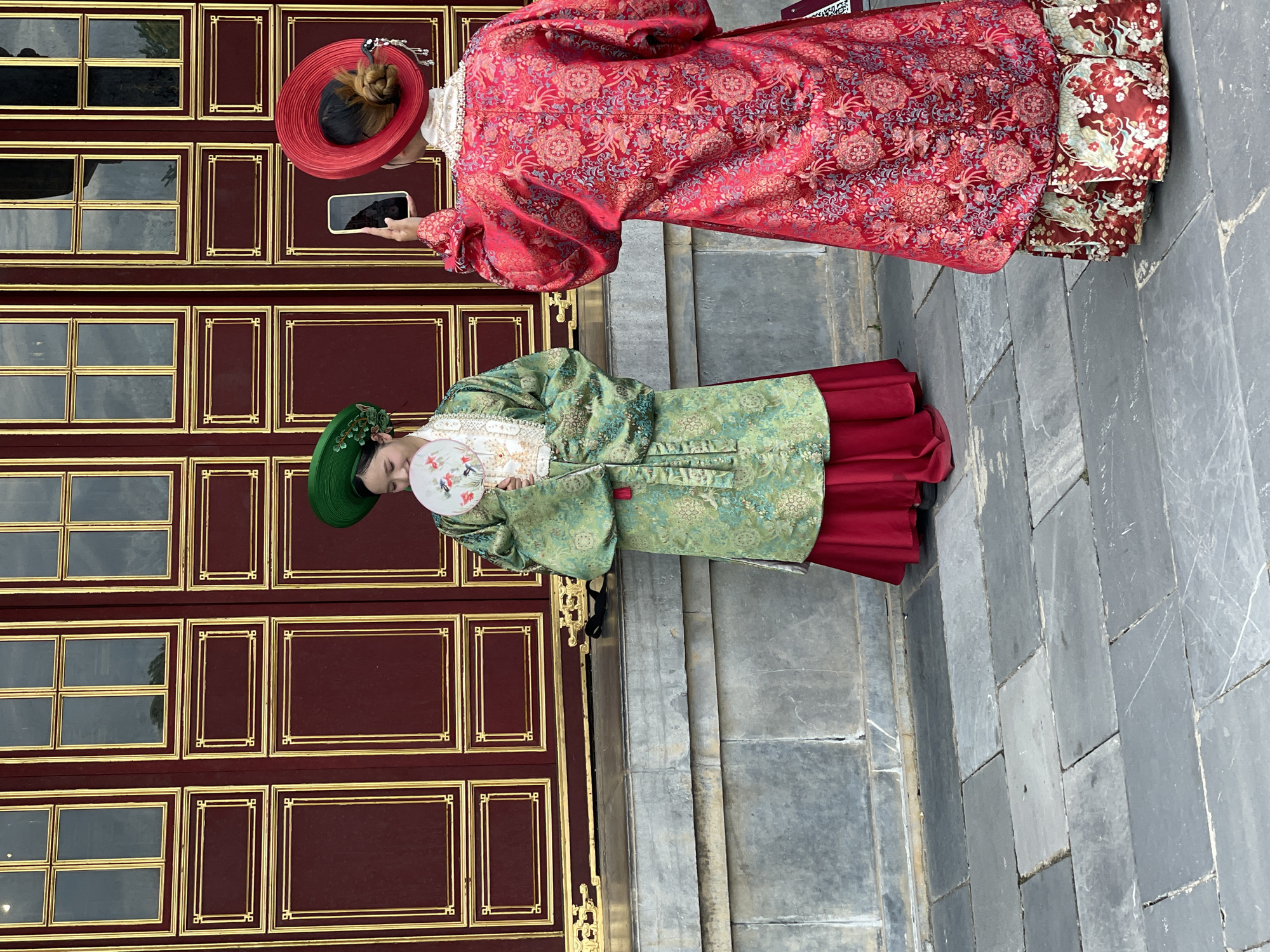 Two individuals are positioned in front of a large, ornate red door with gold trim. The individual on the left wears a green and gold patterned robe over a red skirt and a green conical hat, holding a fan. The individual on the right faces away from the camera, wearing a vibrant red robe with intricate patterns over a patterned skirt and a red hat. They are holding a smartphone, seemingly taking a photo. The ground is paved with large, flat gray stones. The architectural style suggests a historic or traditional setting, possibly within the Imperial City of Huế, located in Phú Xuân Ward, Vietnam.
