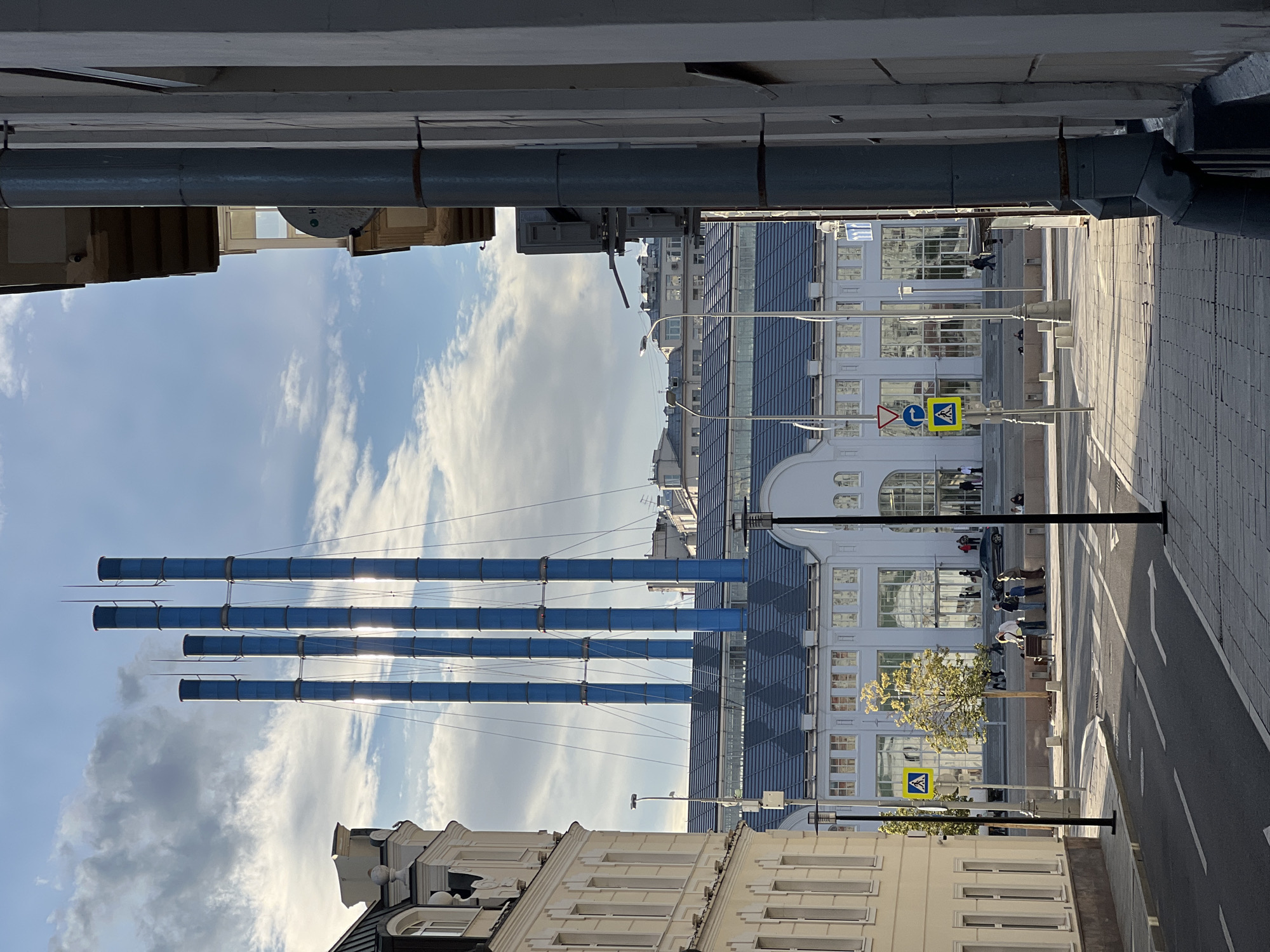This media file captures an outdoor urban scene, rotated 90 degrees clockwise. The foreground is dominated by the lower sections of white and beige buildings with multiple windows, typical of historical European architecture. Above these, a large, gray, horizontal pipe structure extends across the frame, casting shadows.
The midground features a bright sky filled with white, wispy clouds. Several tall, narrow, blue cylindrical structures, possibly part of an art installation or industrial equipment, rise vertically against the sky. Sunlight glints off these pipes. Further back, a modern white building with a dark blue, sloped roofline and numerous windows is visible. A tree with sparse, light green leaves stands in front of it. A paved street runs parallel to the buildings, marked with white lane lines and a pedestrian crossing.
Several people are visible, mostly small figures in the distance, walking on the sidewalk and near the street. Some are gathered near the tree and on benches. Traffic lights and road signs, including a yellow pedestrian crossing sign, are present. The lighting suggests it is daytime, likely late afternoon given the long shadows. The overall mood is one of everyday urban life bathed in natural light. There is no visible text in the image.