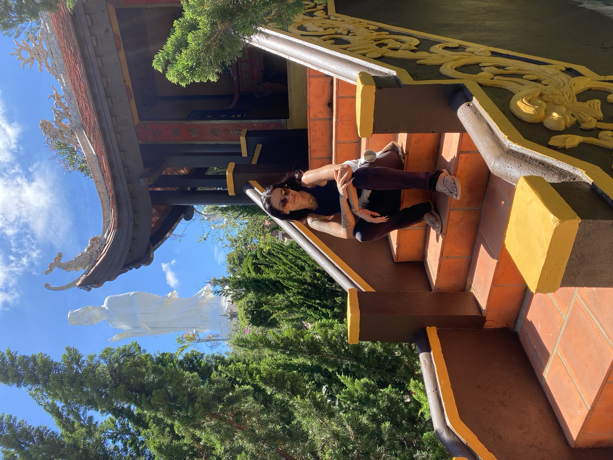 A woman with dark hair and sunglasses sits on an outdoor staircase in front of a temple. She is wearing a black tank top, dark pants, and light-colored sandals. The staircase is made of terracotta tiles and has a dark brown railing with yellow accents. Behind her, a large white statue of a woman, possibly a deity, stands tall against a bright blue sky with white clouds. The temple building has a traditional East Asian architectural style with a red tiled roof and ornate decorations. Green trees and foliage surround the temple and statue. The scene appears to be in a Buddhist temple in Thôn 5, Vietnam.