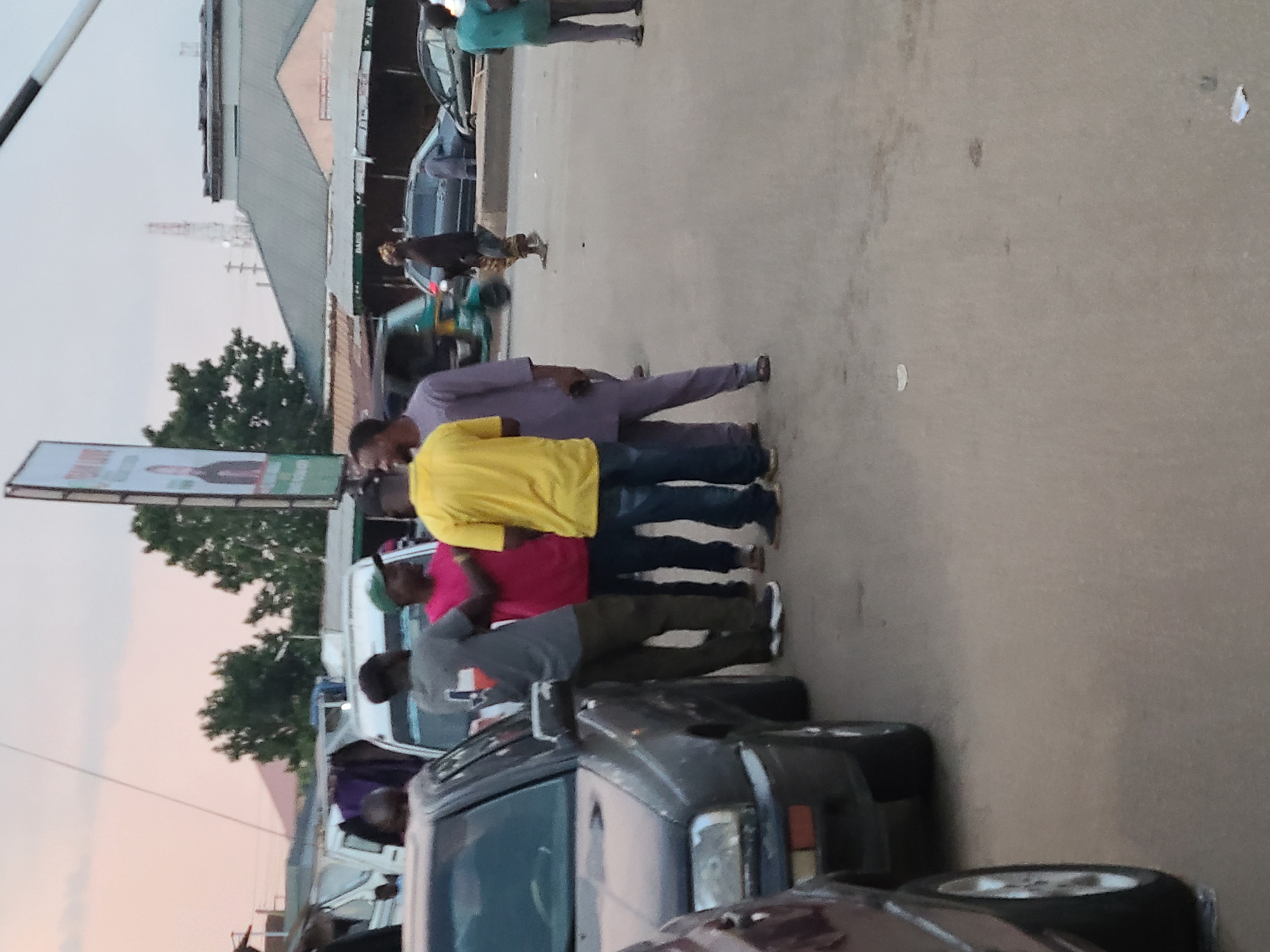 A group of men are gathered on a paved street, likely a marketplace or busy road in Jos, Nigeria, based on the presence of a billboard advertising a political candidate and the general urban infrastructure visible. In the foreground, a dark-colored sedan is parked on the left, with its front driver's side visible. Behind it, another dark-colored car is partially obscured. A group of five men are in discussion or interacting in the center of the frame. One man in a gray t-shirt with a Texas flag design on the back is facing away from the camera. Next to him, a man in a bright pink t-shirt and dark pants is also facing away. A man in a yellow t-shirt and jeans is turned to his right, engaged in conversation with the others. Another man in a light purple traditional outfit is standing to his left, holding a phone. A fifth man, wearing a dark outfit, is standing slightly behind and to the right of the group. Further in the background, several white vans and cars are parked or moving. A multi-story building with a gray roof and windows is visible, along with a large billboard featuring a picture of a man. The sky appears overcast with a hint of pink, suggesting either dawn or dusk. The ground is asphalt, with some scattered debris.