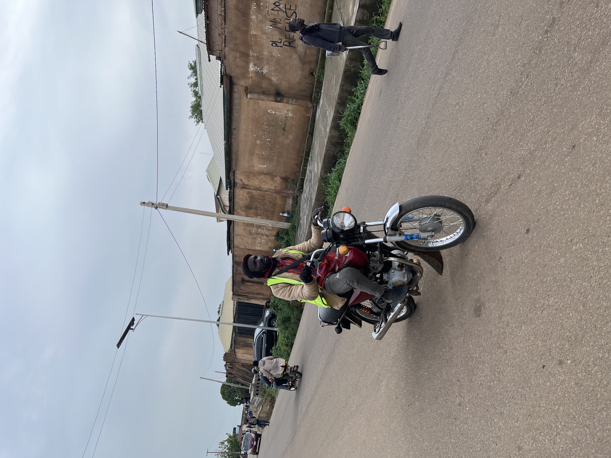 A man rides a motorcycle down a paved road in Gura Top, Nigeria. He is wearing a tan jacket over a red and black plaid shirt, a yellow reflective vest, and a black beanie. A red bag is slung across his shoulder. He has a serious expression on his face. The motorcycle is dark red with chrome accents and a silver fender. In the background, there is a tan building with graffiti on the wall that reads "PL. NO REFUSE". Green vegetation lines the side of the road. The sky is overcast, suggesting it is a cloudy day. Further down the road, another motorcycle with two riders is visible, as well as several cars and more pedestrians. A streetlight is mounted on a pole in the foreground. The overall scene depicts a moment of daily life and transportation in a Nigerian town.