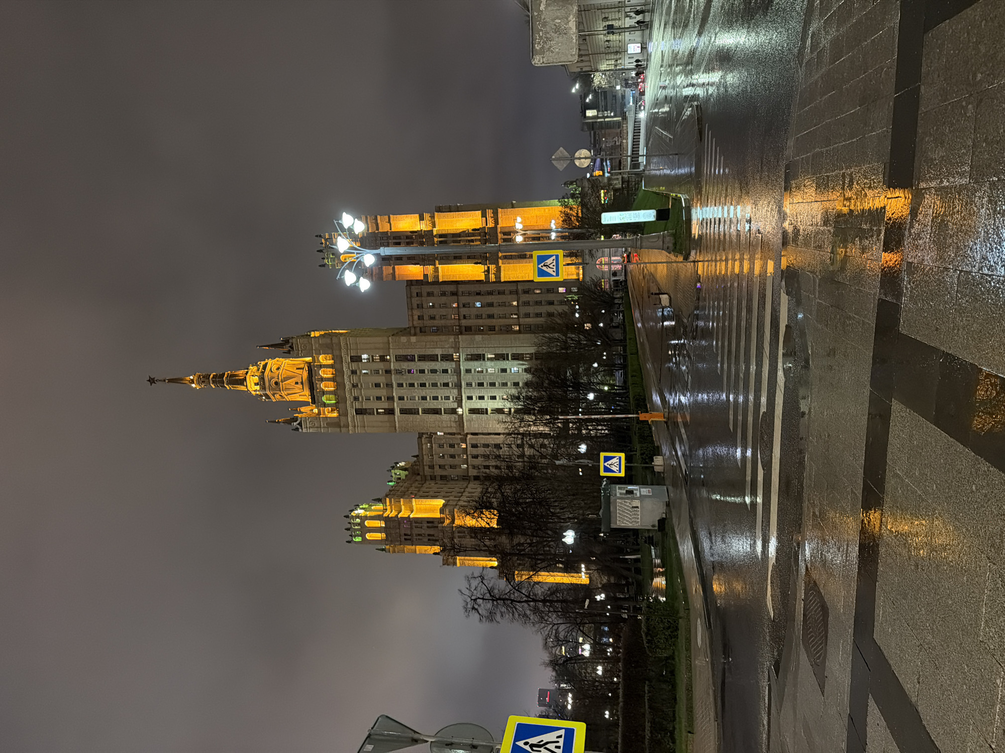 The image depicts a nighttime scene in Moscow, Russia, likely after a rain shower, as evidenced by the wet pavement and reflections of light. The prominent structure is one of the Seven Sisters, a group of Stalinist skyscrapers. This building is brightly lit, with a golden glow emphasizing its architectural details. In the foreground, there's a pedestrian crossing with white stripes and wet surfaces, reflecting the surrounding lights. Sidewalks are also visible, with streetlights adding to the overall illumination. A pedestrian crossing sign is also visible. The sky is overcast, contributing to the somber yet vibrant atmosphere of the urban setting. The building is a skyscraper, composed of many different segments, with lights illuminating the architectural details. The roads are empty.