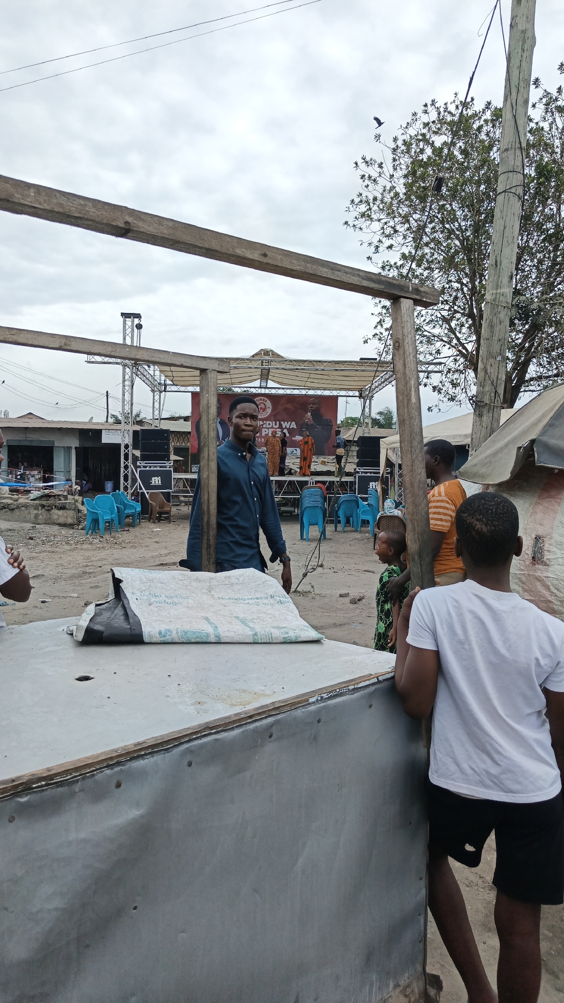 This image depicts an outdoor scene, likely in Dar es Salaam, Tanzania, featuring a temporary stage setup for an event. In the foreground, a metallic-surfaced table or stand is partially visible, covered with a large white bag featuring text in a foreign language. Several individuals are present. A man in a dark blue shirt stands near the center, facing the camera. To his right, a group of people are gathered, including a child in a green and black patterned shirt and an adult in an orange and white striped shirt. A young person wearing a white t-shirt and black shorts is visible from behind, leaning on the stand. In the background, a stage is set up with speakers and a large banner displaying images and text, suggesting a political or promotional event. The ground appears to be unpaved, and several blue plastic chairs are arranged in front of the stage. The sky is overcast, and a large tree with visible branches is to the right of the frame, near a utility pole. Buildings with corrugated metal roofs are visible in the distance.